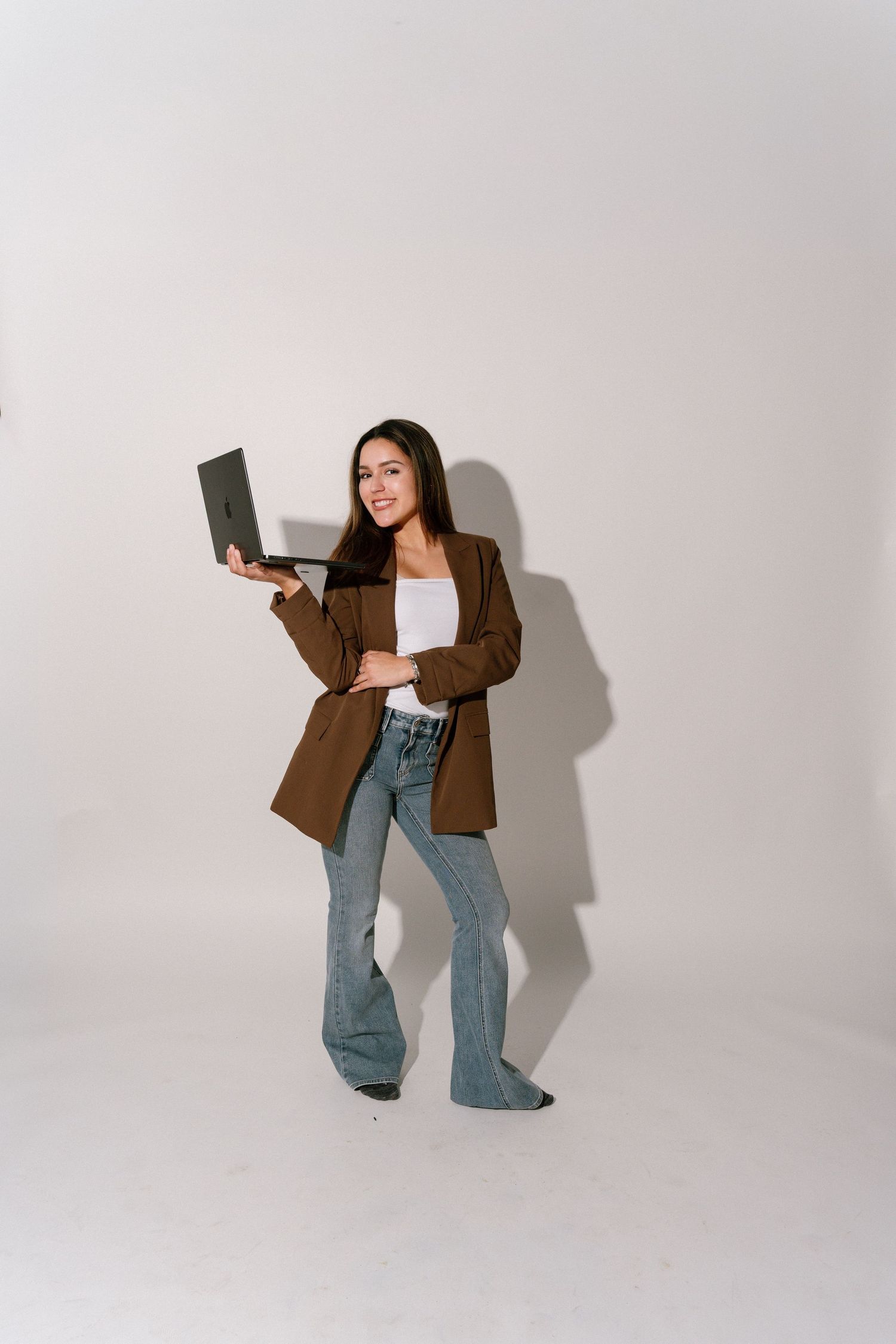 Young woman in brown blazer and flare jeans holding a laptop, posing confidently against a white background.