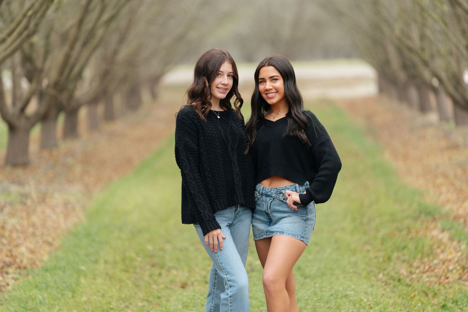 Two friends in casual outfits pose together on a path between rows of bare trees.