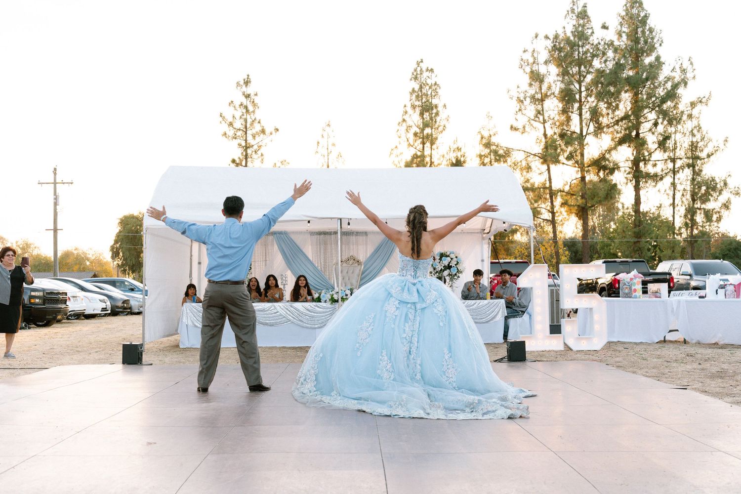 A couple celebrates at an outdoor quinceañera with a light blue ballgown against a white tent at sunset.