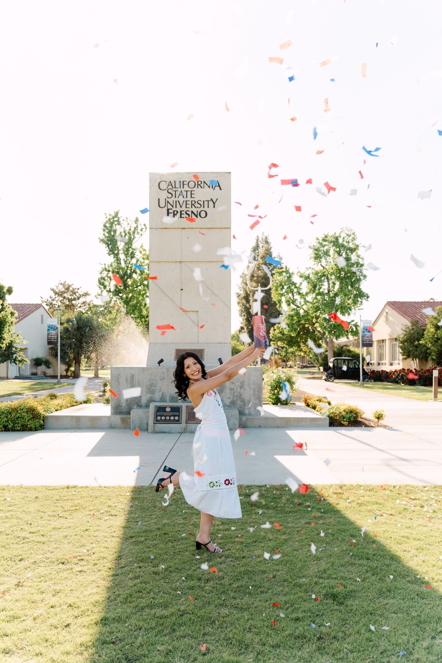 Graduate celebrates in front of California State University Fresno sign with colorful confetti flying in the air.