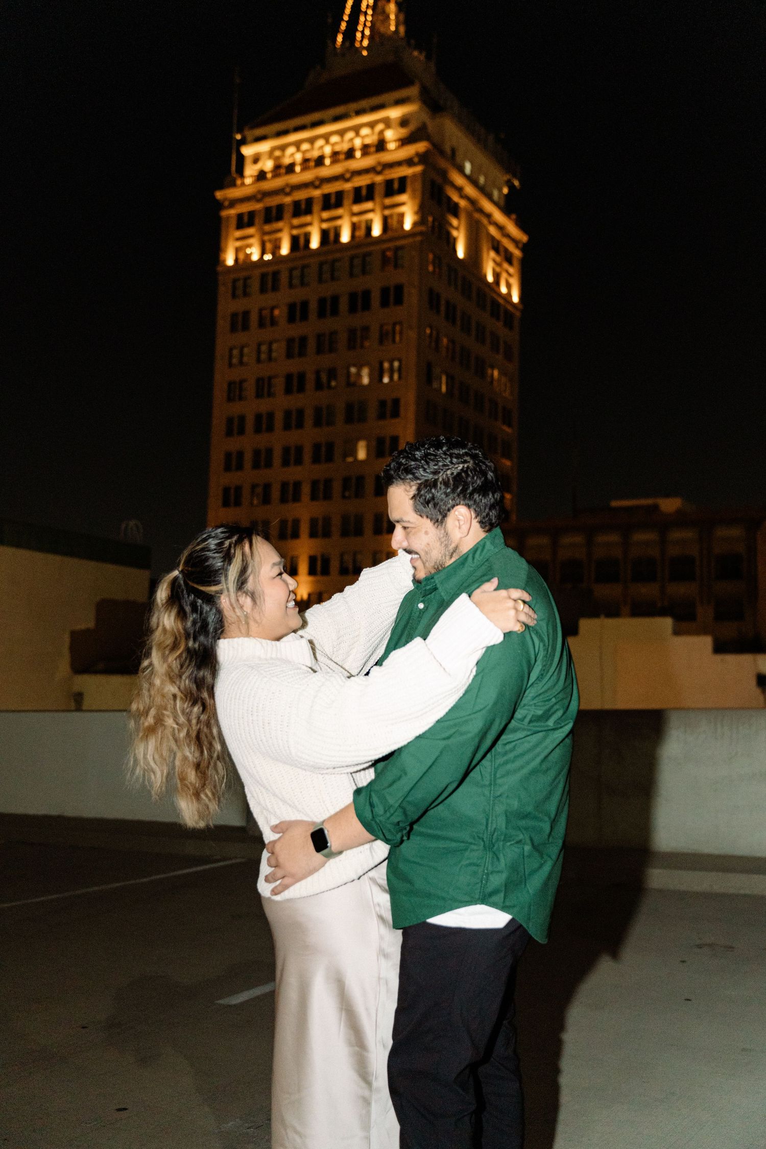 A couple dances together on a rooftop at night with an illuminated building in the background.