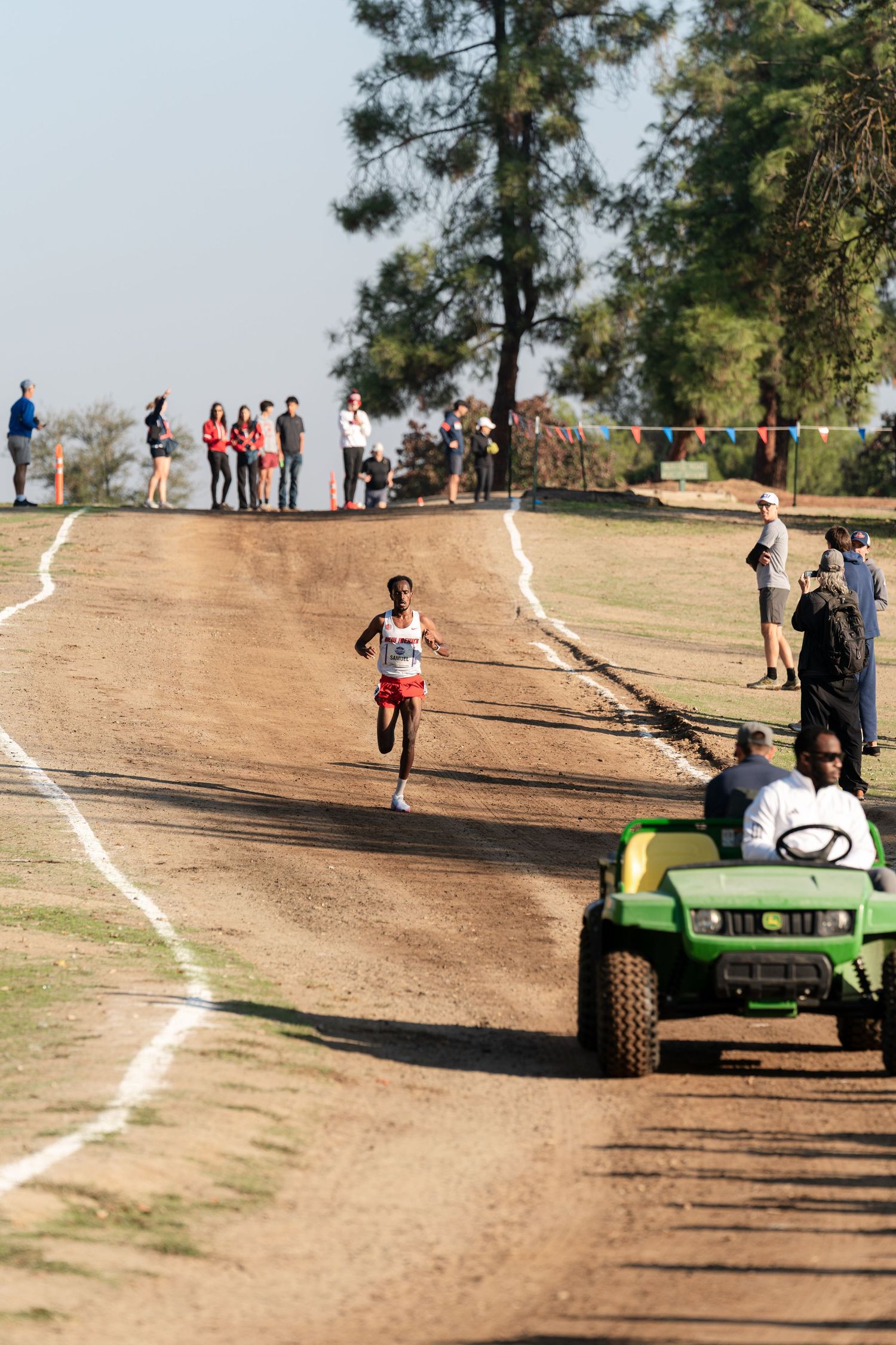 Runner competing on dirt cross country course with spectators and golf cart along marked path during race event.