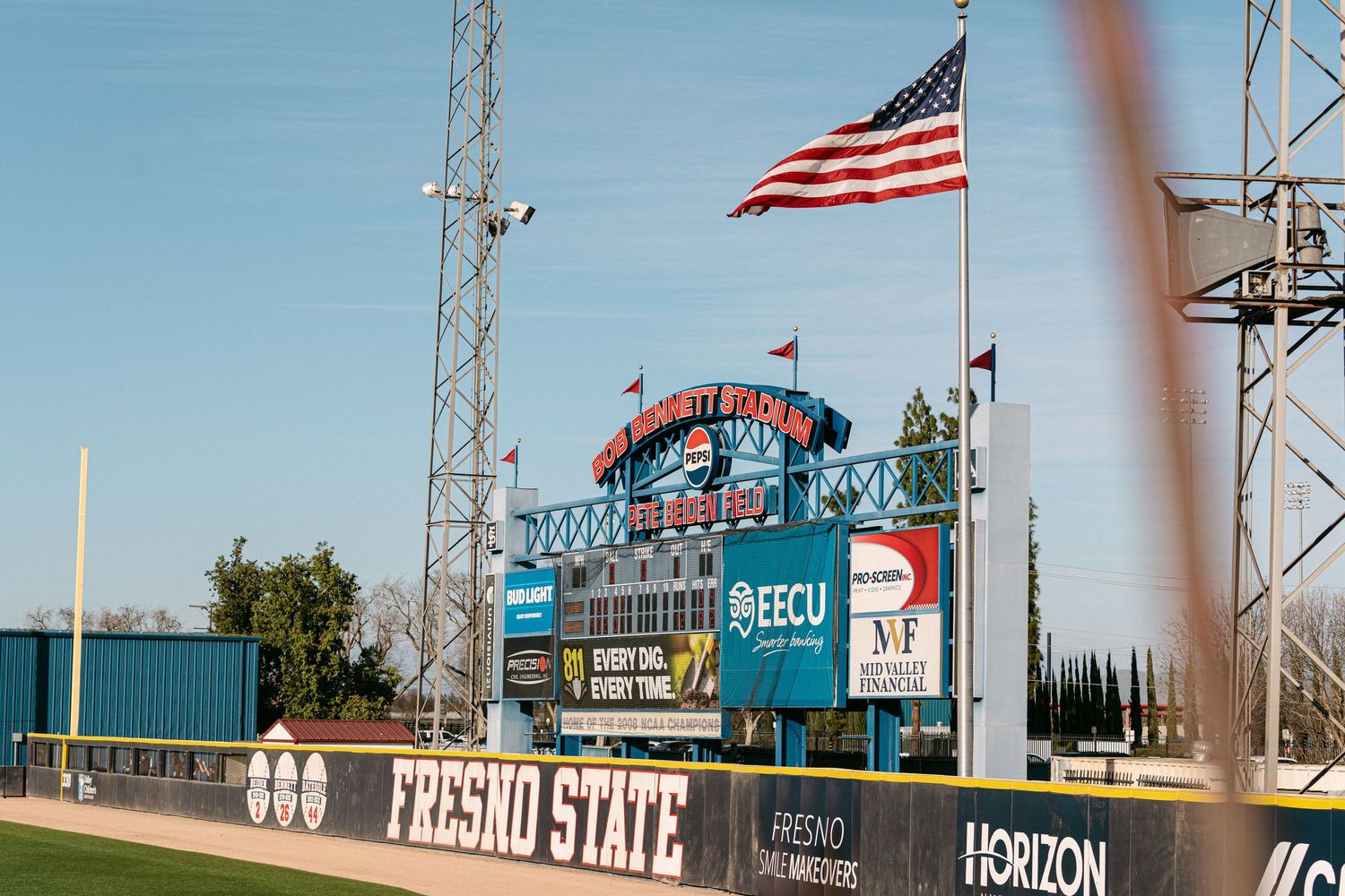American flag waves atop a communications tower against a bright blue sky with buildings visible in background.