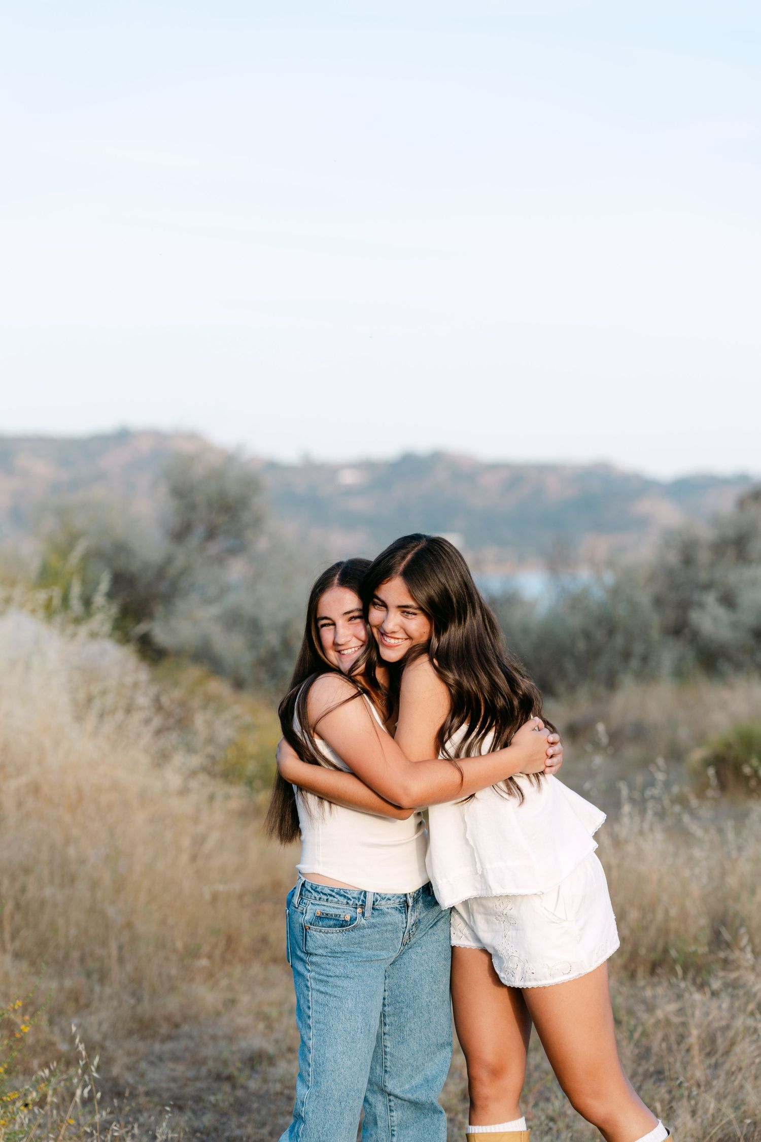 Two young women hugging and smiling outdoors with hills and blue sky in the background.