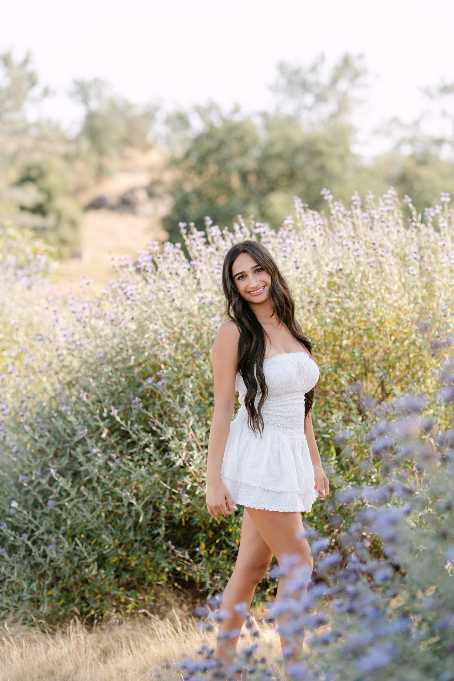 Young woman in white strapless mini dress smiling among purple wildflowers in a sunlit field.