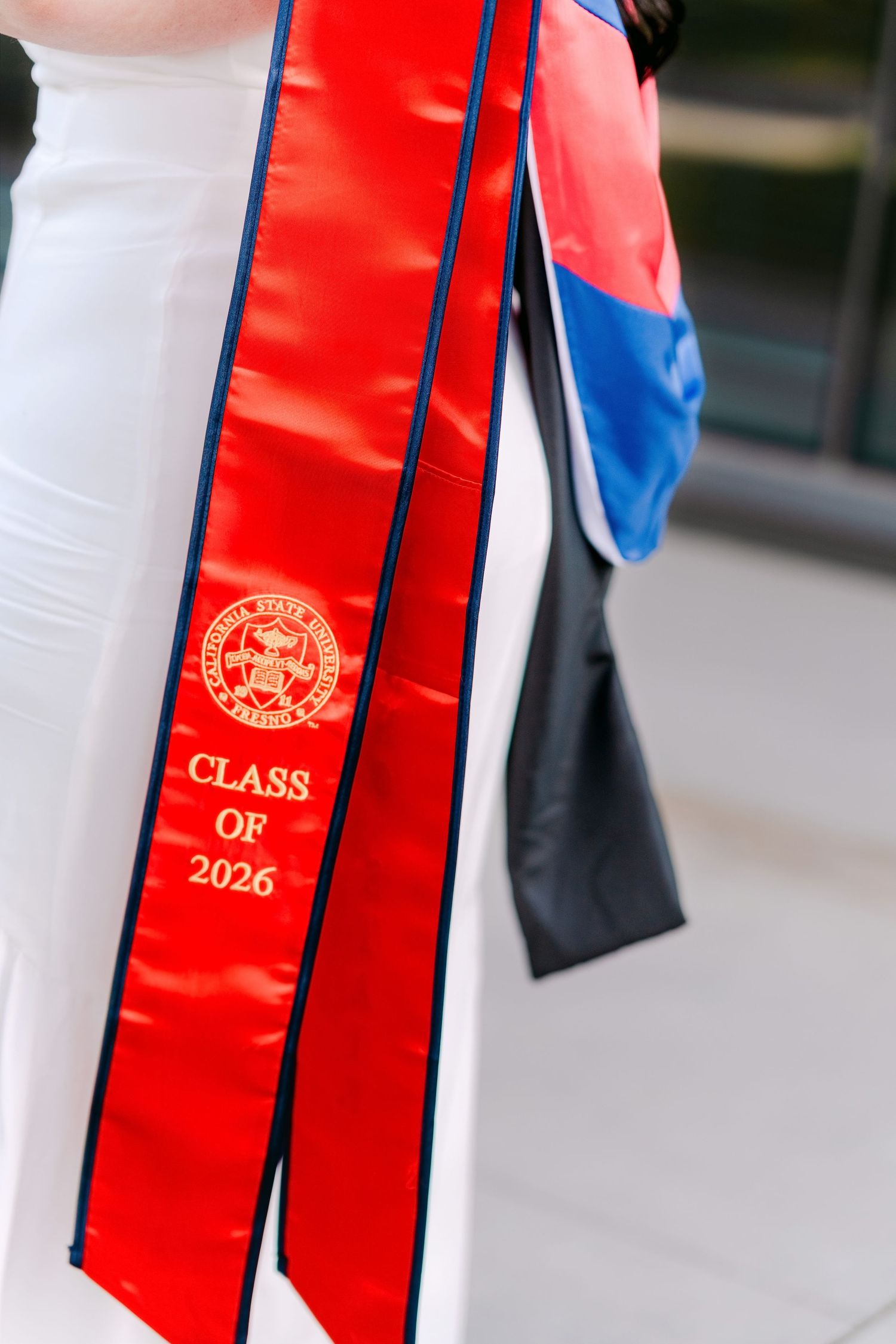 Red graduation sash with gold emblem reading Class of 2026, worn over white graduation gown.