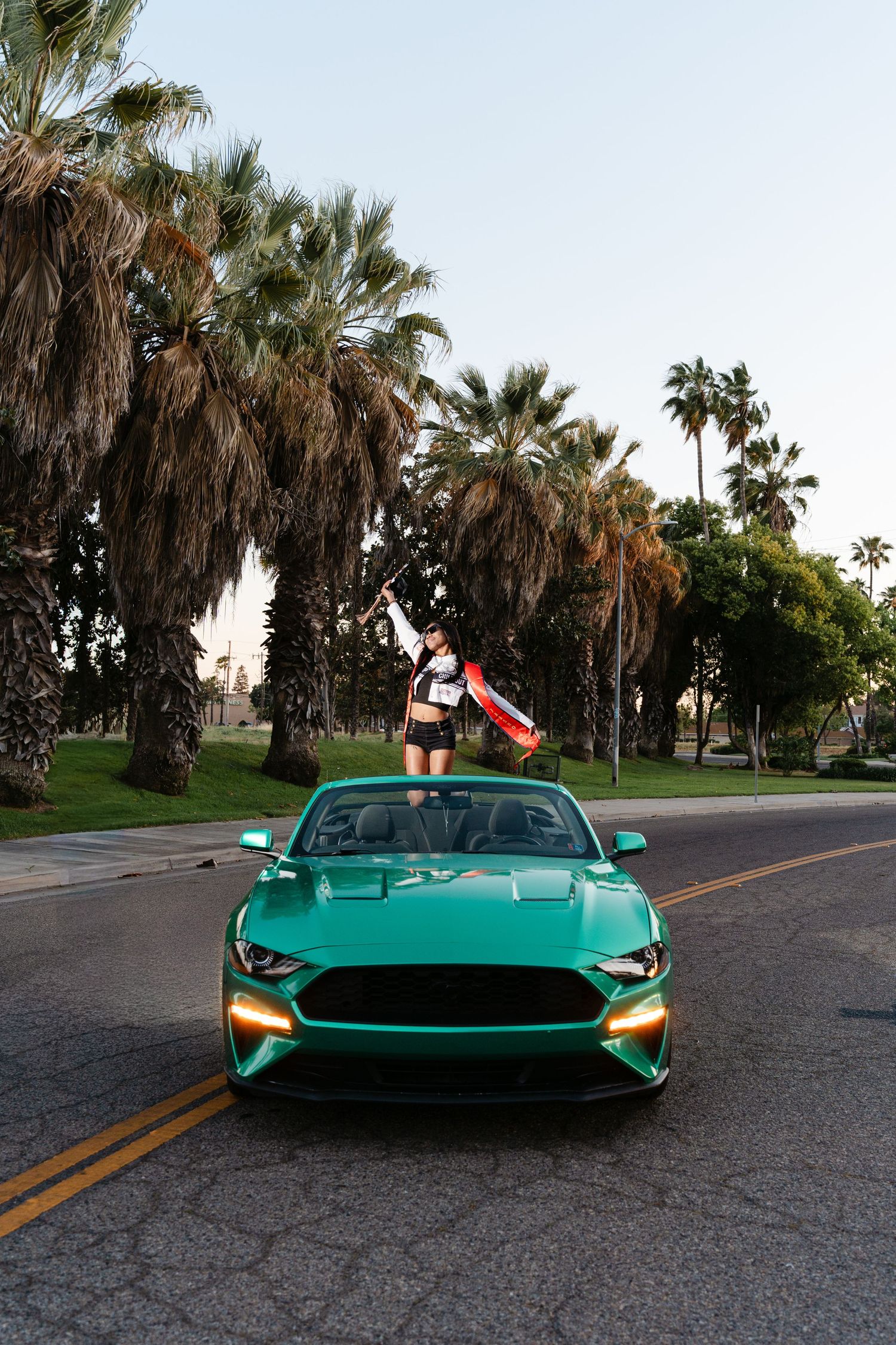 A woman stands on a green Ford Mustang convertible holding a flag, surrounded by palm trees on a sunny street.