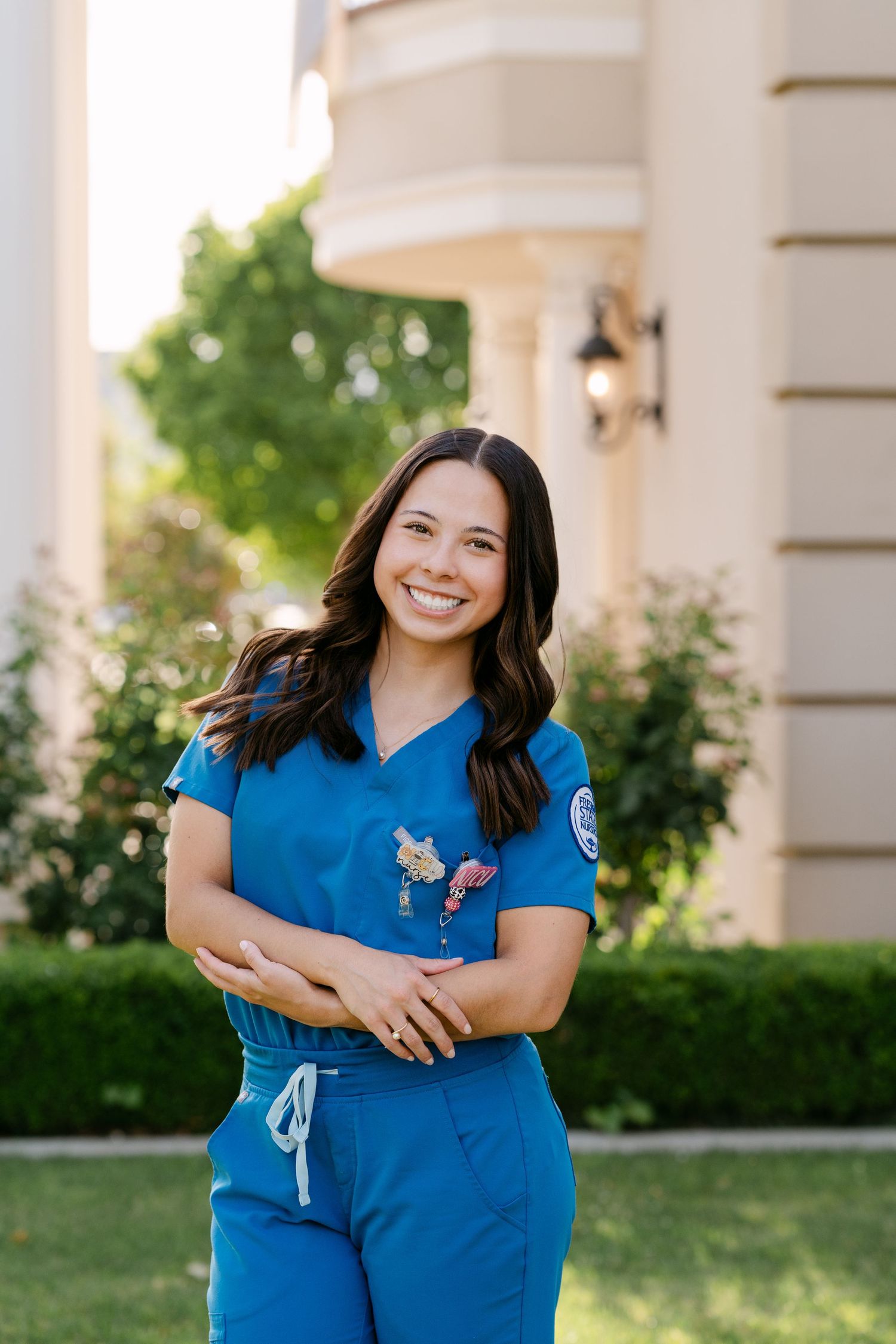 Smiling young woman in blue medical scrubs with badge posing outdoors near a building with green trees.