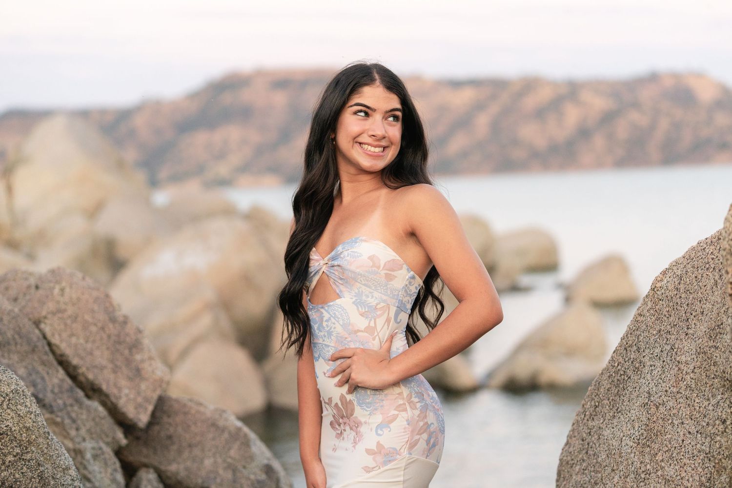 A woman in a floral dress poses against a rocky coastal backdrop during golden hour.