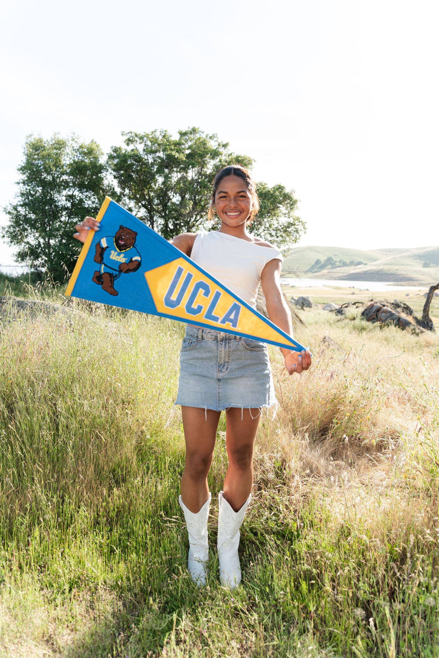 Smiling young woman holding a UCLA pennant flag outdoors in a grassy field, wearing a denim skirt and white boots.