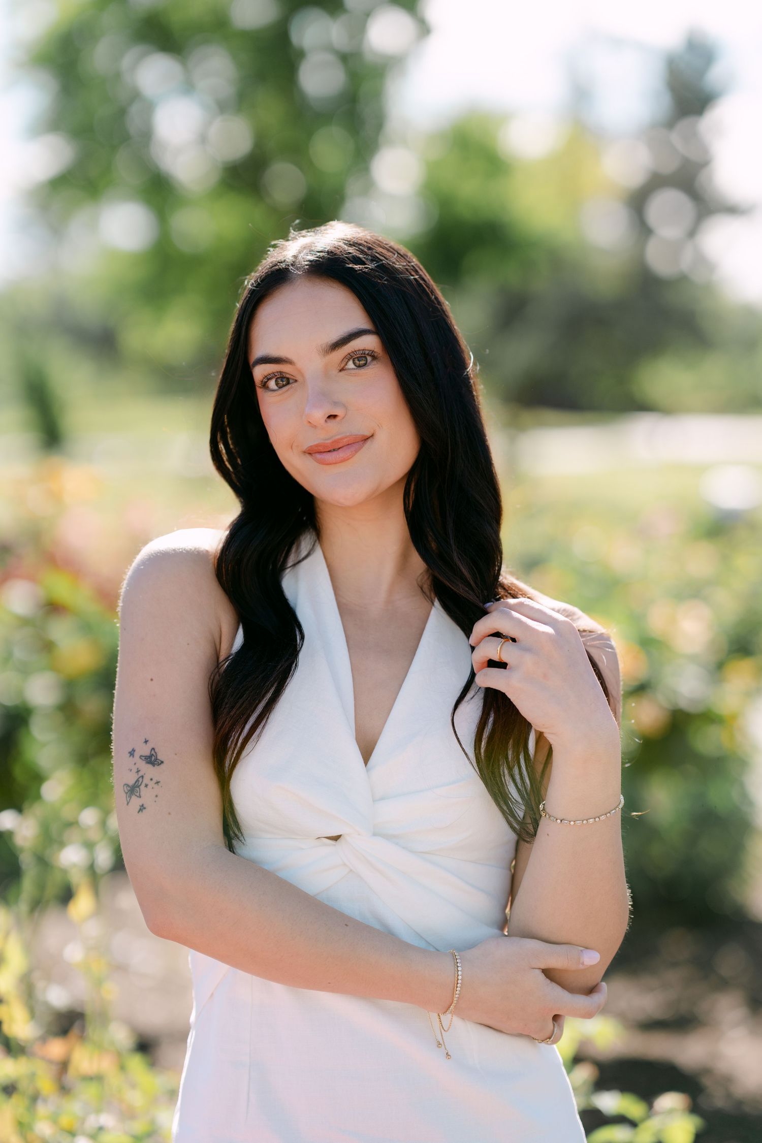 Young woman with dark hair in white dress, floral tattoo on arm, posing outdoors in a sunny garden setting.