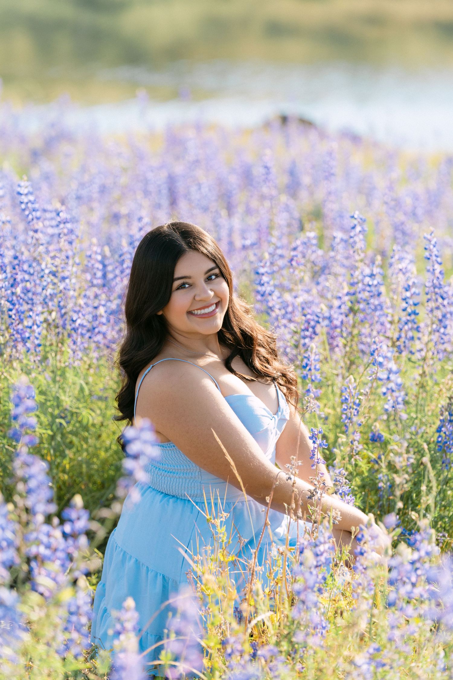 Smiling young woman in a light blue dress standing in a vibrant purple lupine flower field near a lake.