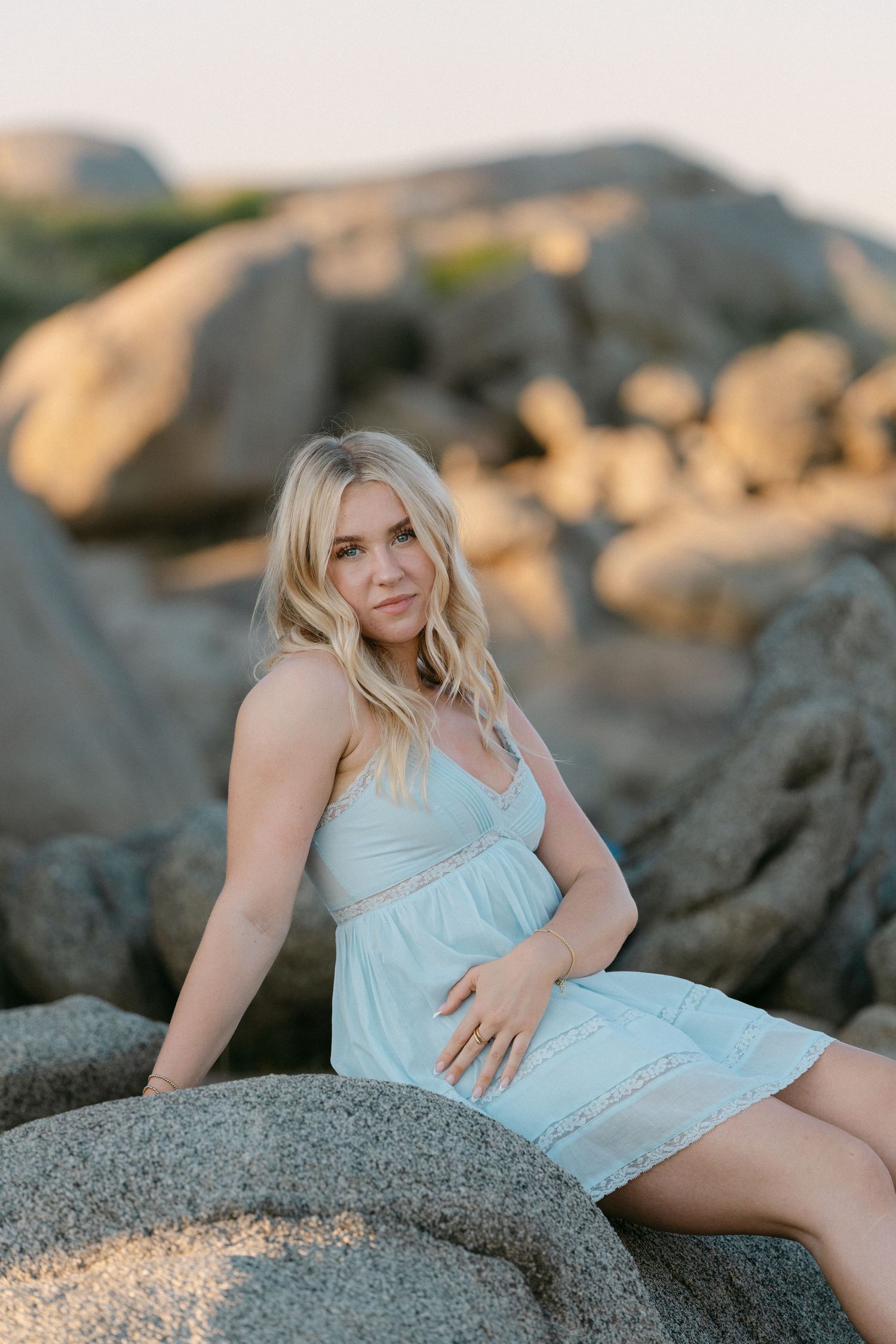 Pregnant woman in light blue dress sitting on coastal rocks at golden hour, with wavy blonde hair.