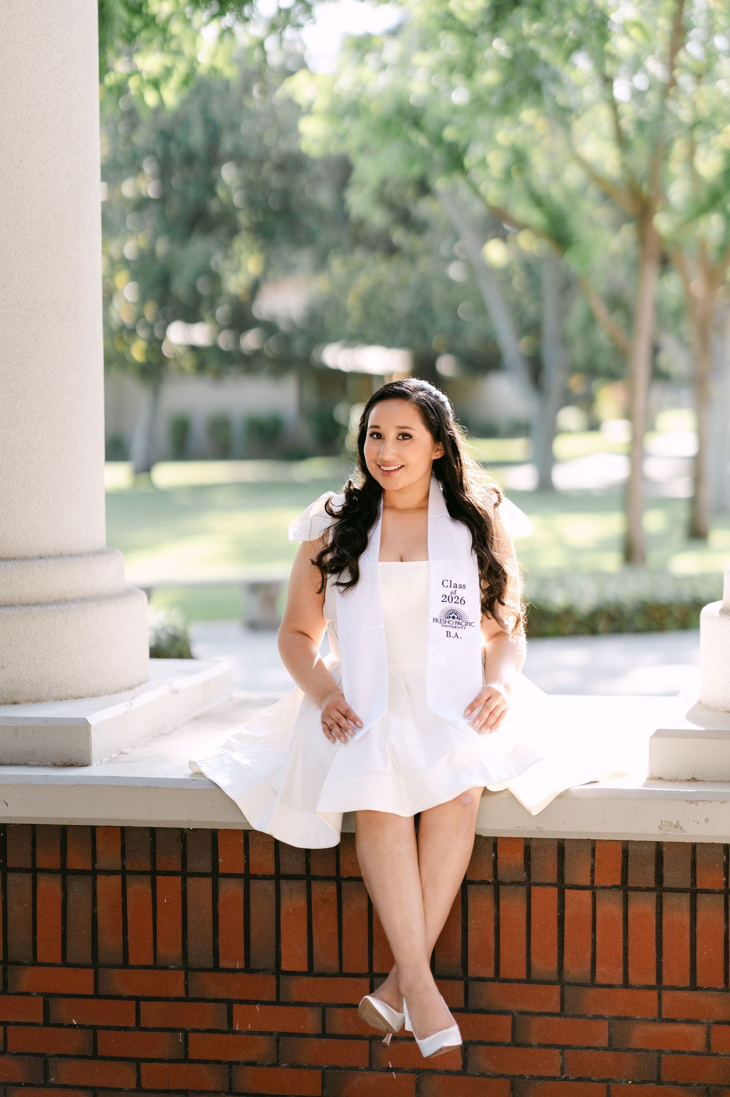 Young woman in white dress and graduation stole sitting on brick wall outdoors on sunny day.