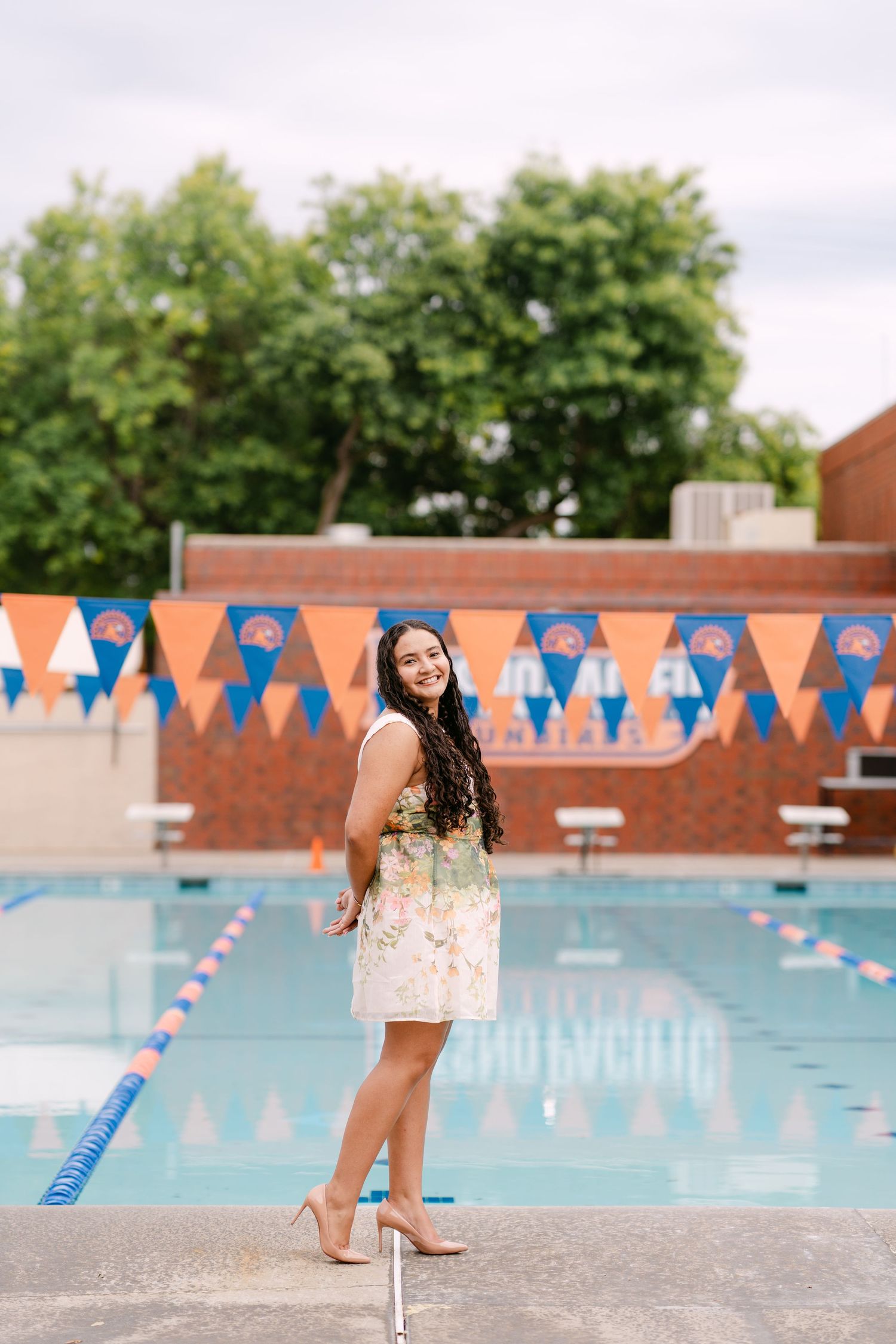 Young woman in floral dress and heels posing poolside with orange and blue pennant banners.