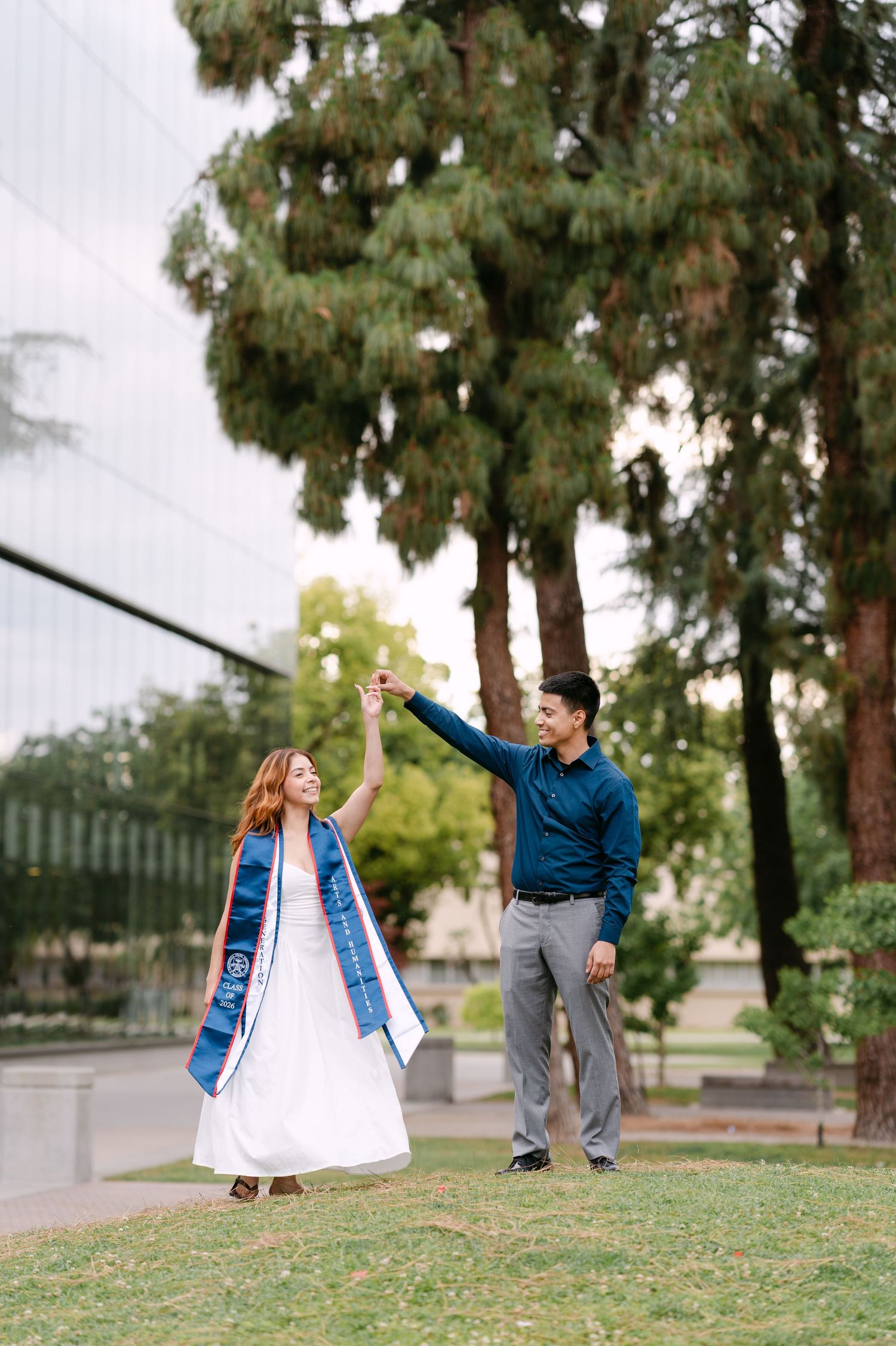 Graduate in white dress and blue stole dances with man in teal shirt on green campus lawn.