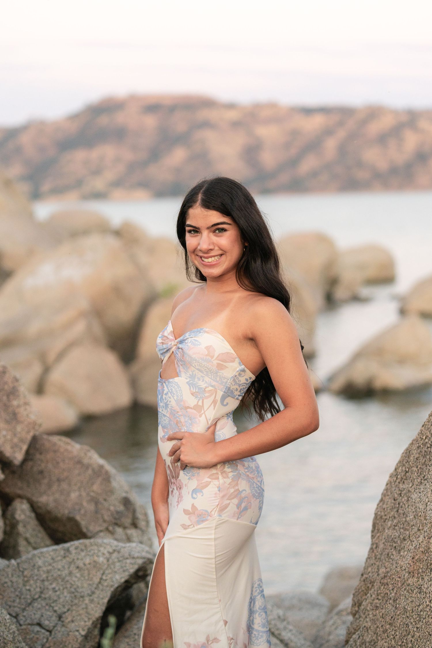 A person in a white lace dress poses gracefully against coastal rocks during a seaside photoshoot at sunset.