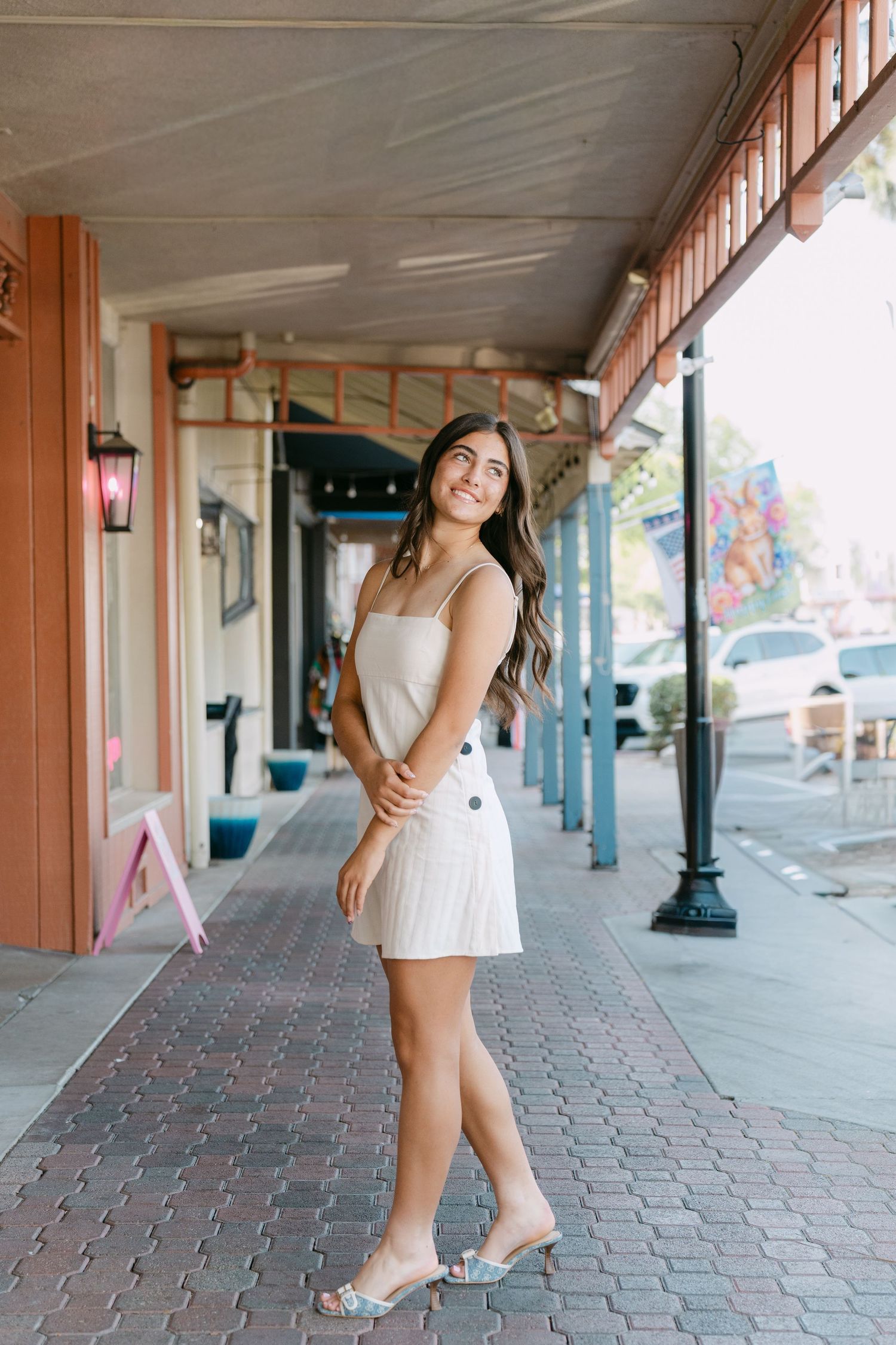 A person in a white summer dress poses along a covered walkway with brick pavement and wooden pillars.