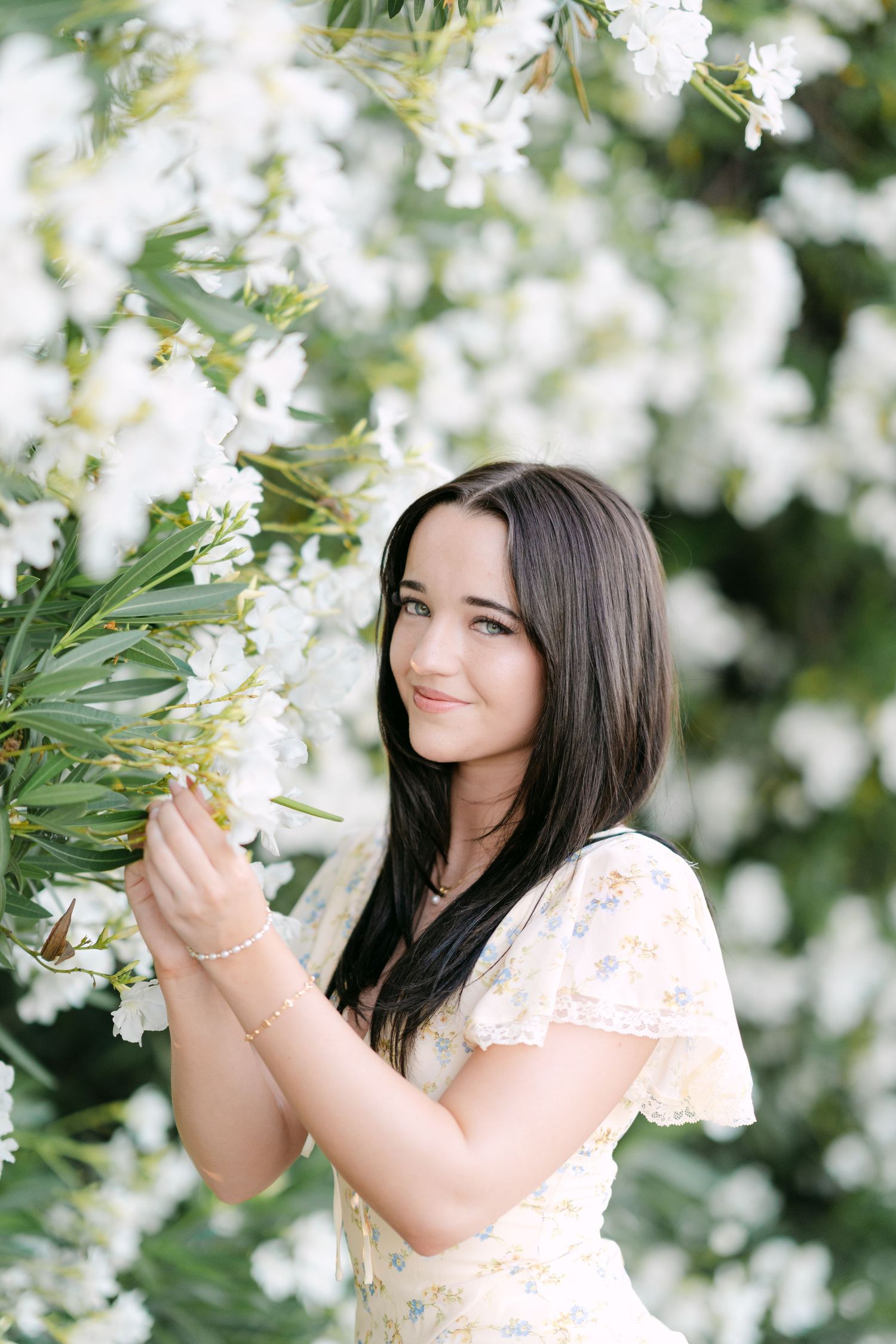 Young woman in floral dress holding white blooms, surrounded by flowering shrubs in a garden setting.