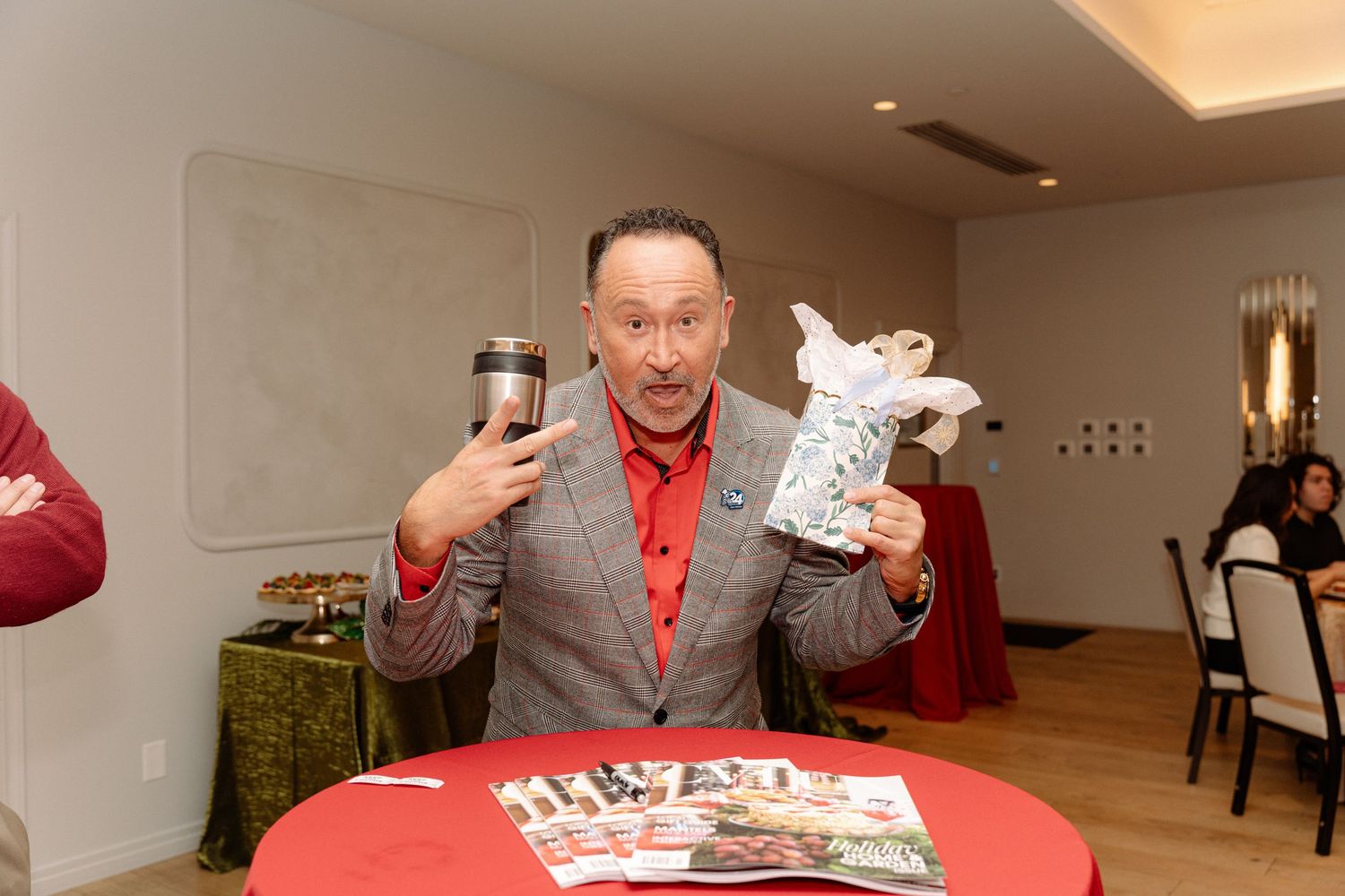 Man in gray suit and red shirt excitedly holds up a phone and stuffed animal at a decorated table.