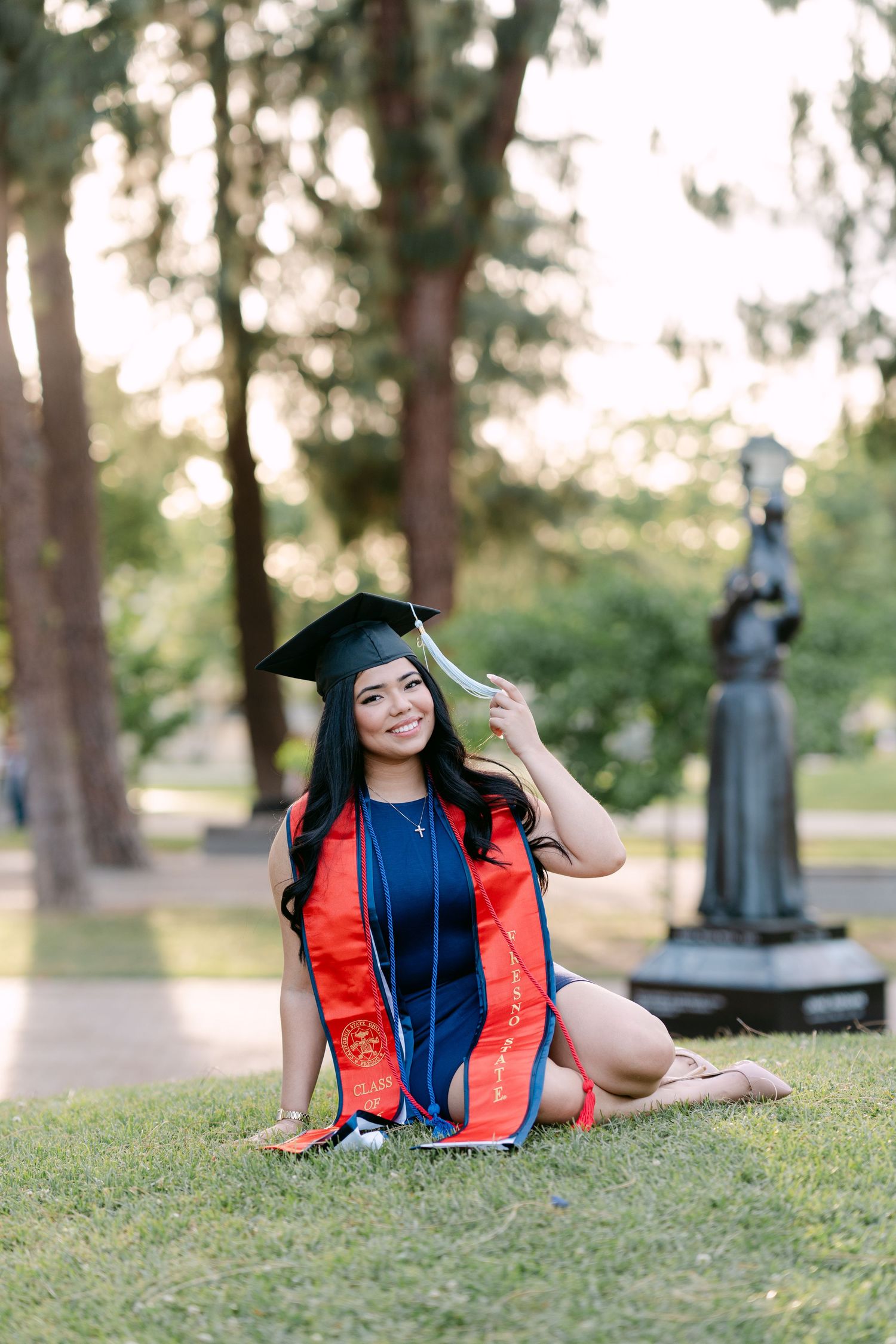 Smiling graduate in cap and gown sitting on grass outdoors, holding tassel, wearing navy and red stole.