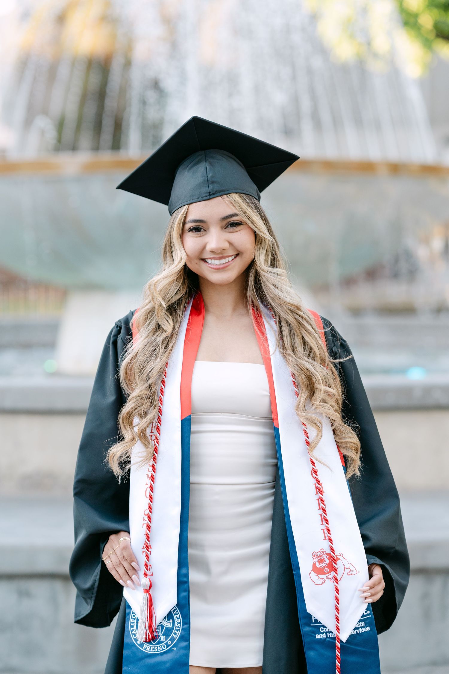 Smiling female graduate in black cap and gown with red and white honor cords, posing in front of a fountain.