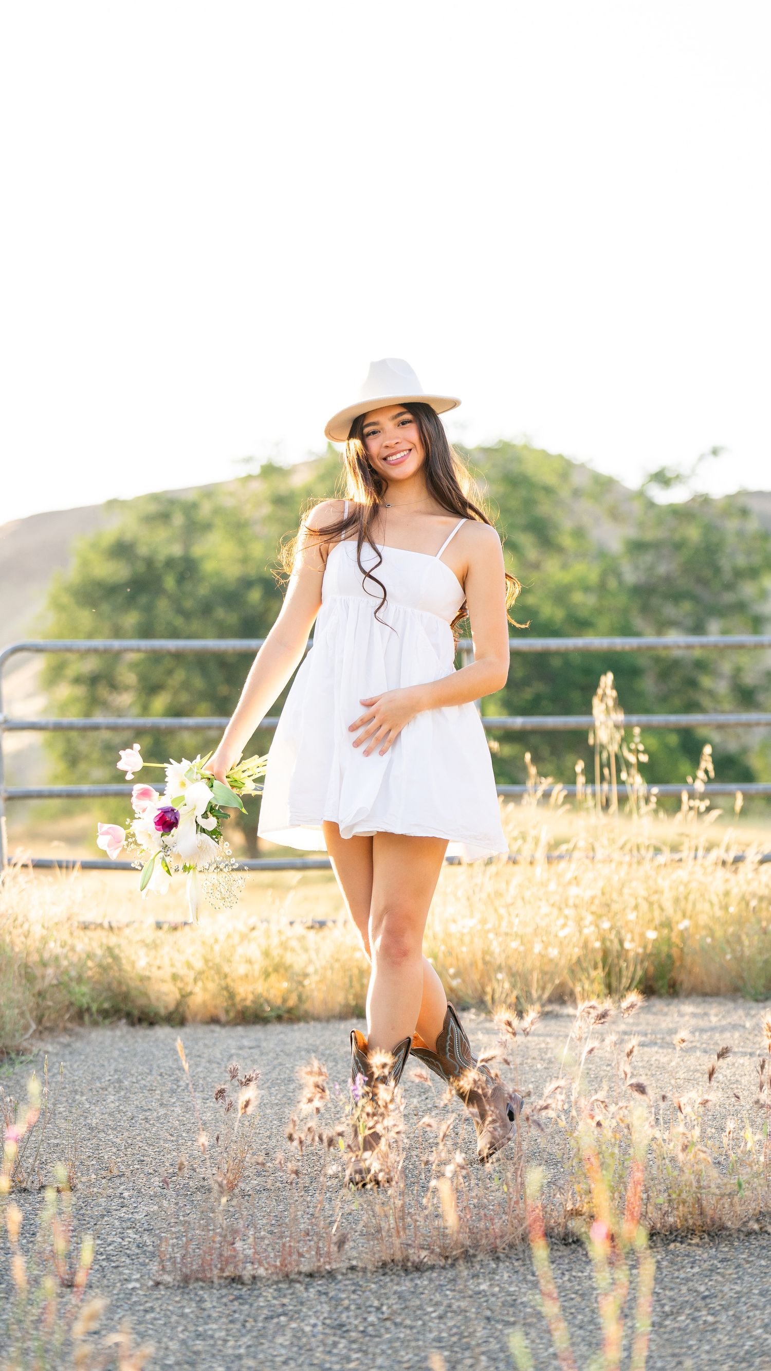 Person in white sundress and hat poses in golden summer field during sunset.