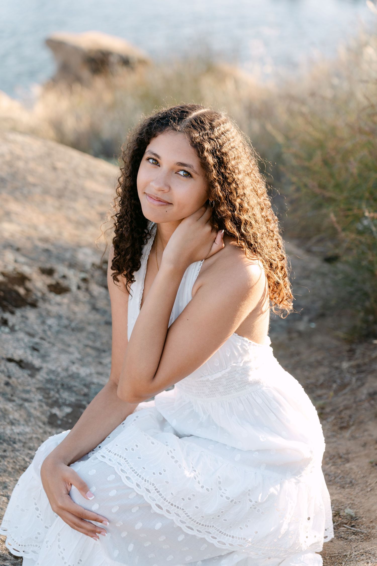Young woman with curly hair in white eyelet dress sitting on rocky shore, smiling softly in golden sunlight.