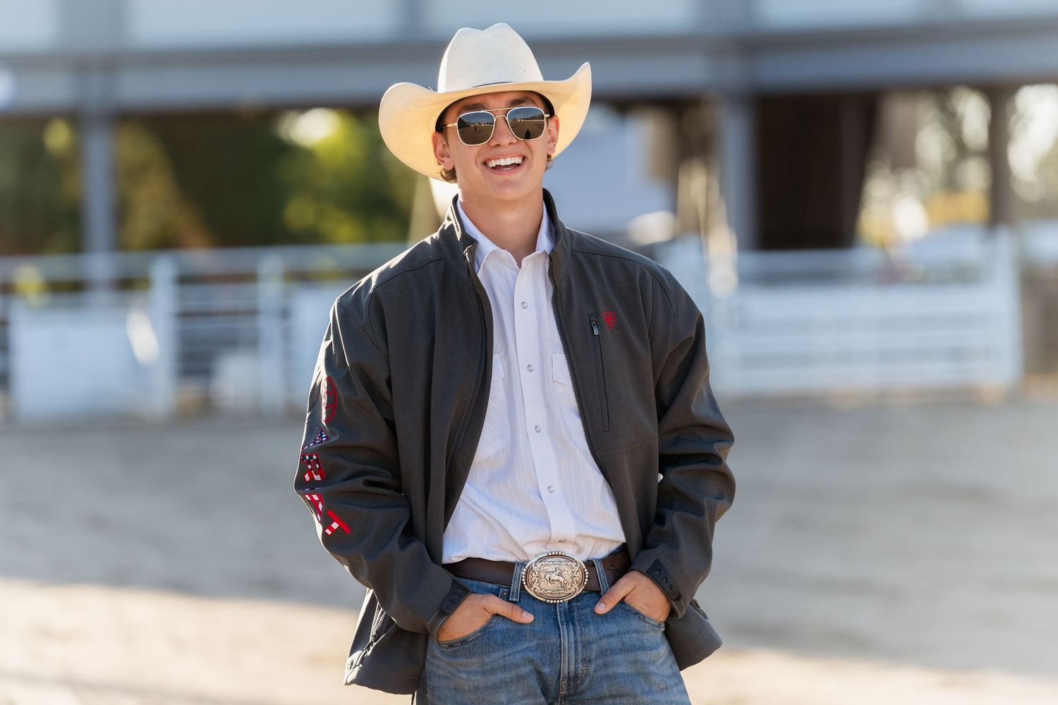 A person in a white cowboy hat and sunglasses smiles while standing outdoors.