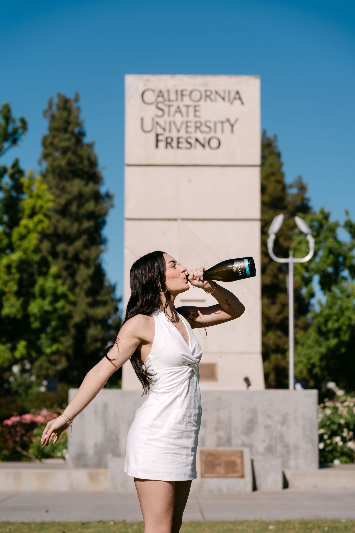 Woman in white dress drinking champagne in front of California State University Fresno sign on sunny day.