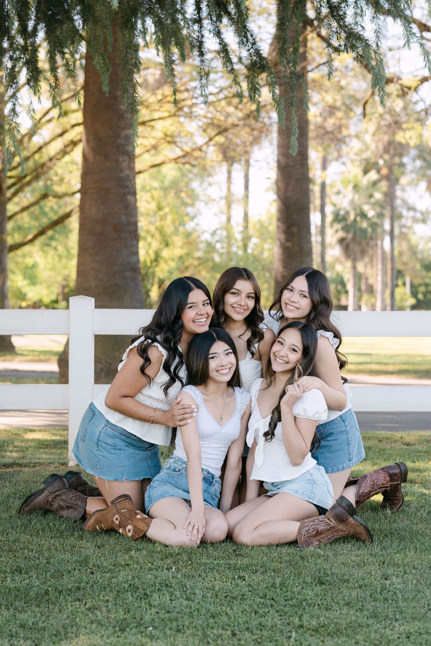 Five young women in white tops, denim skirts, and cowboy boots pose together on green grass near a white fence.