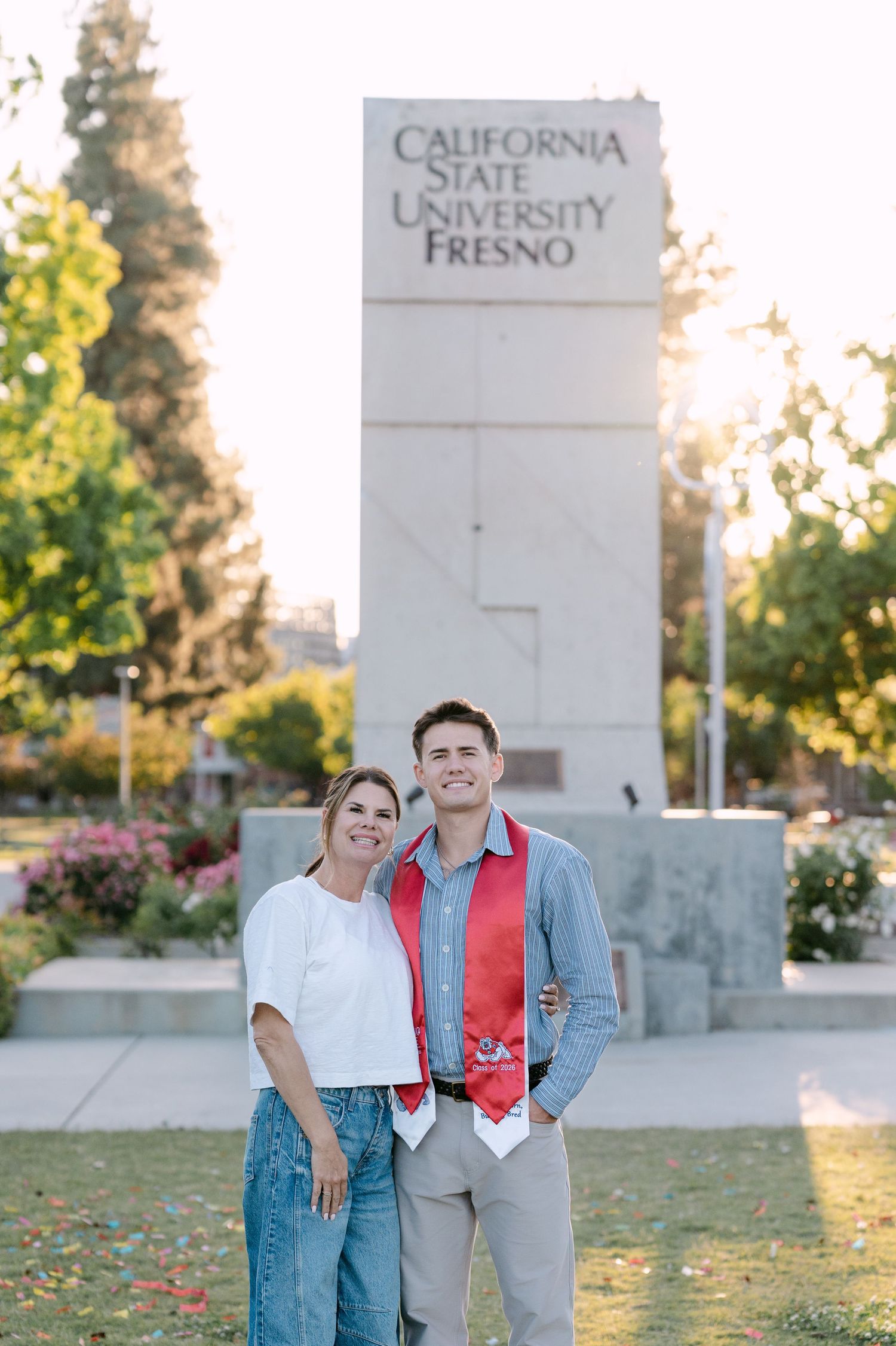 Graduate wearing red stole poses with mother in front of California State University Fresno monument sign.