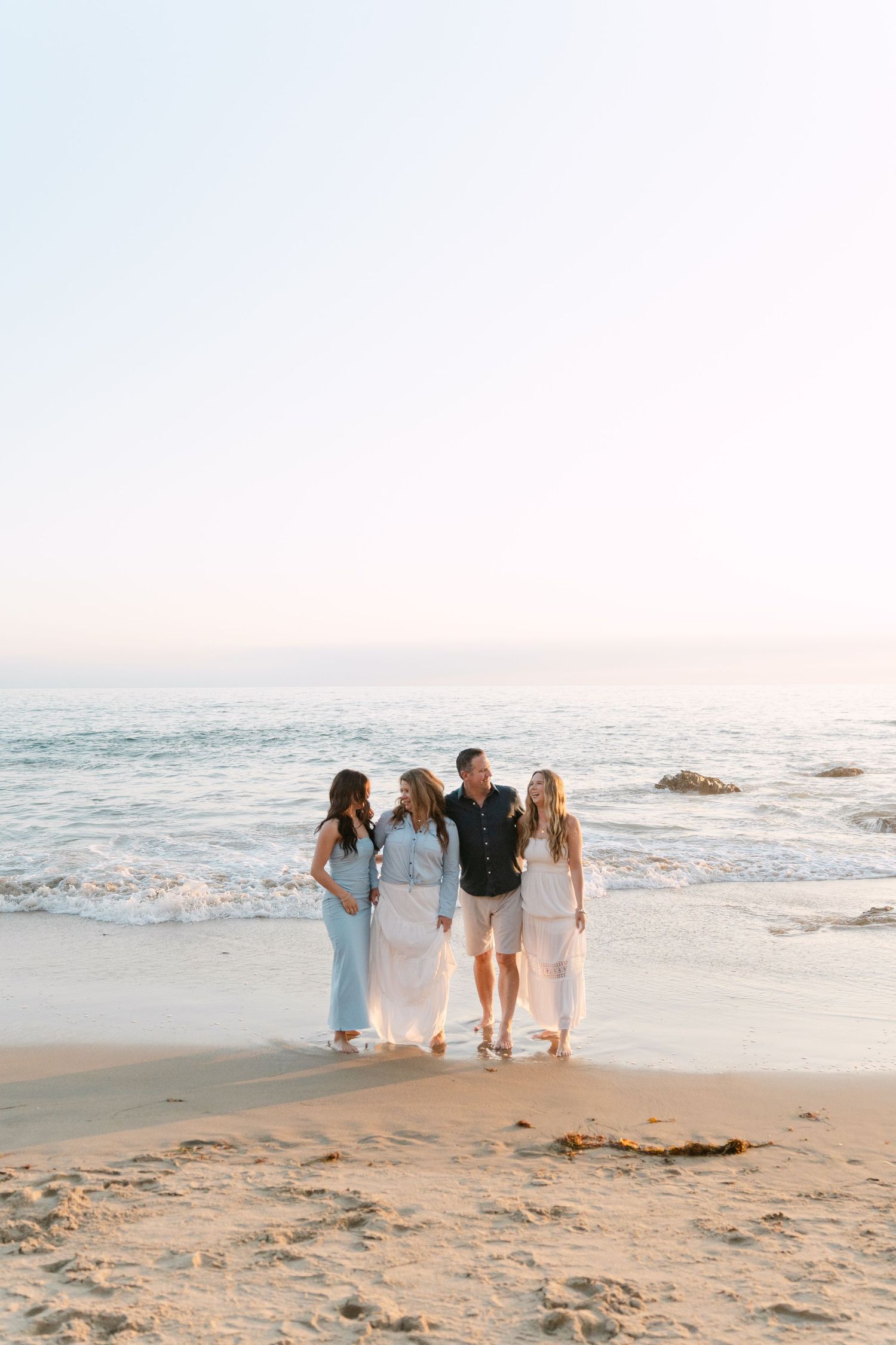 Family of four posing together on a sandy beach at sunset, wearing light casual outfits with ocean waves behind them.