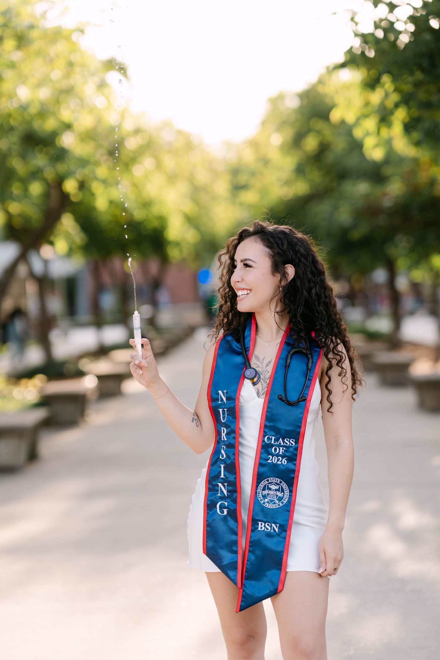 Nursing graduate smiling outdoors in blue BSN Class of 2026 stole, holding a syringe, with tree-lined path background.