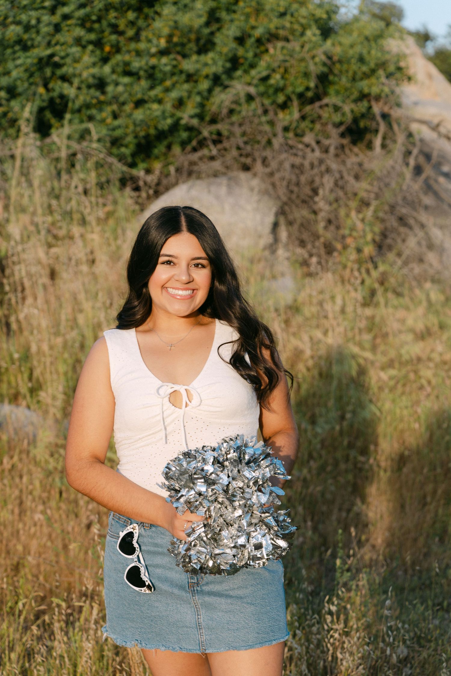 Young woman smiling outdoors holding silver pom-pom, wearing white top and denim skirt in a natural field setting.