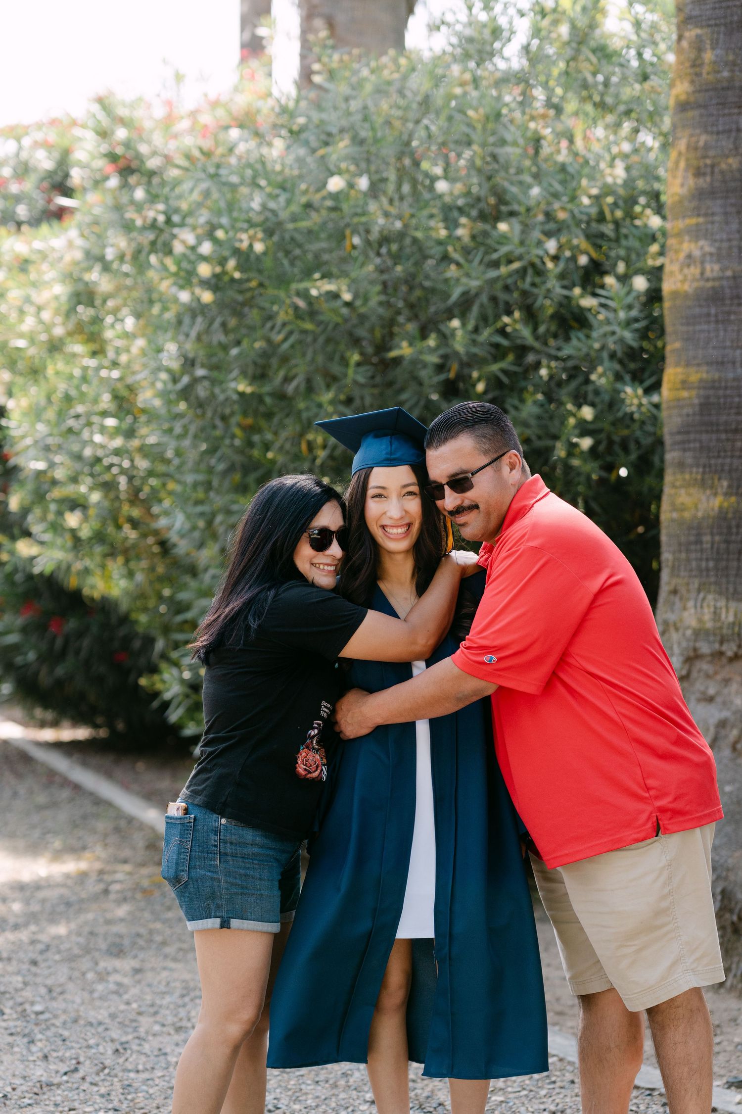 A proud graduate in blue cap and gown poses with smiling parents outdoors amid lush green foliage.
