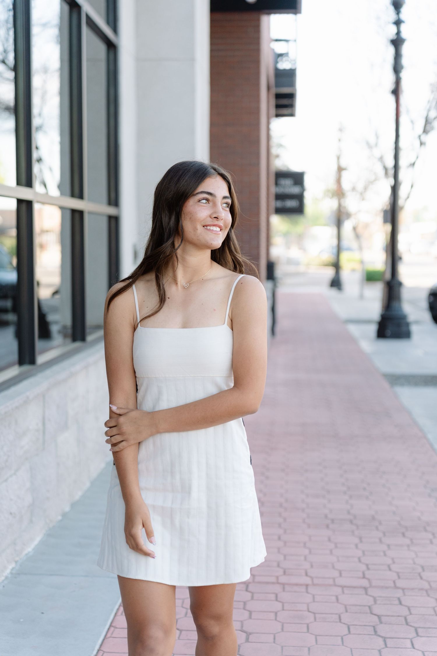 A person wearing a white summer outfit stands on a brick sidewalk near a commercial building.