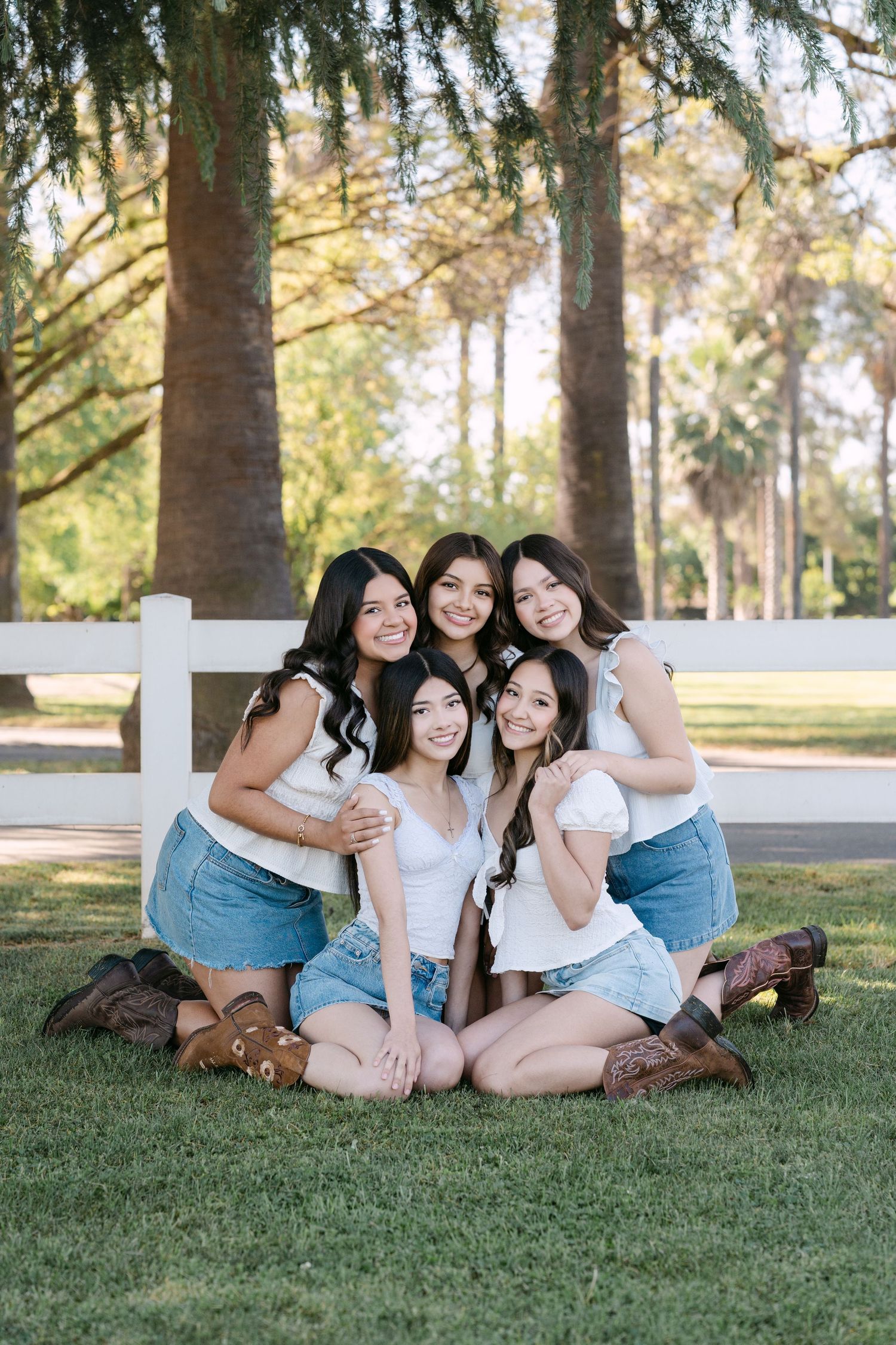 Five young women in white tops, denim skirts, and cowboy boots pose together on grass near a white fence.