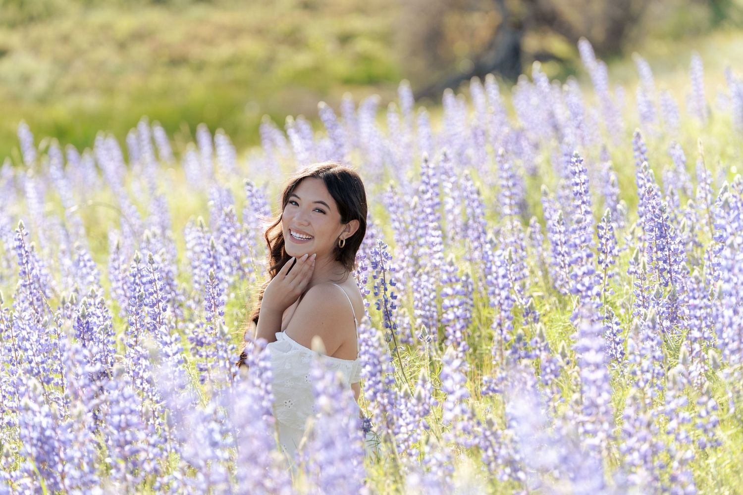 Woman in white dress smiling among purple lupine flowers in a sunny field.