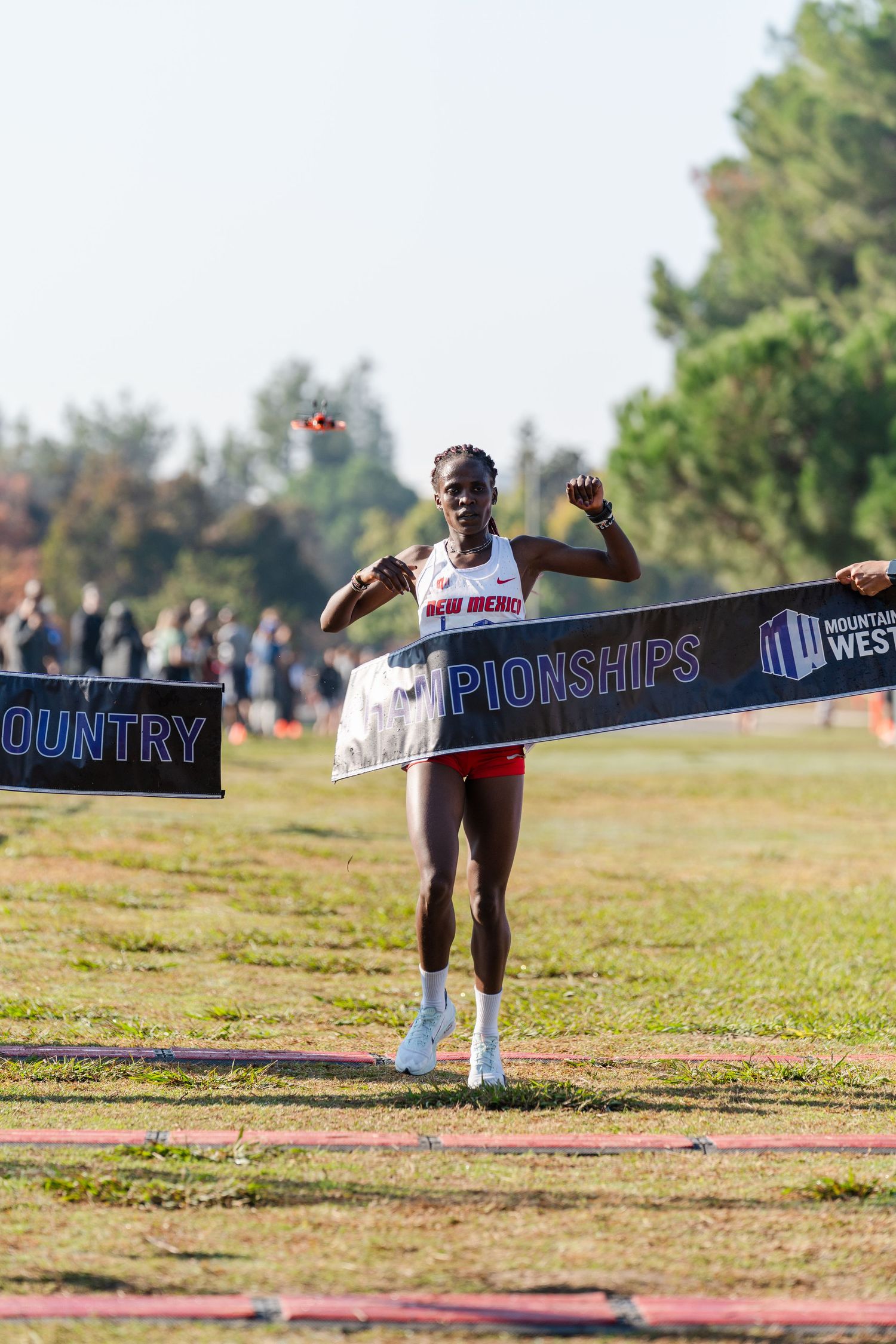 Runner breaks through finish line tape at cross country championships on sunny outdoor course.