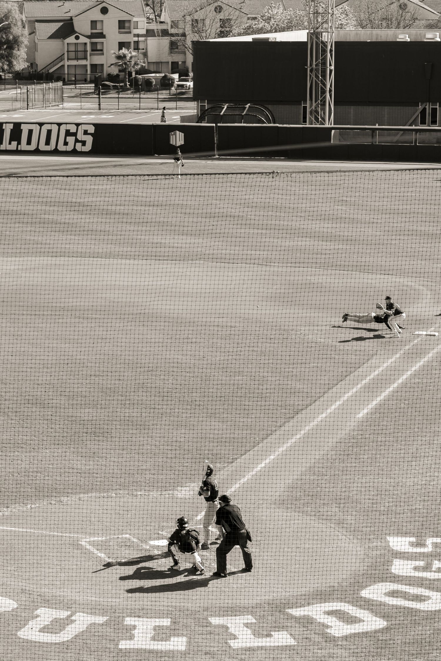 Black and white baseball action shot showing players at home plate during a game with 'BULLDOGS' visible.