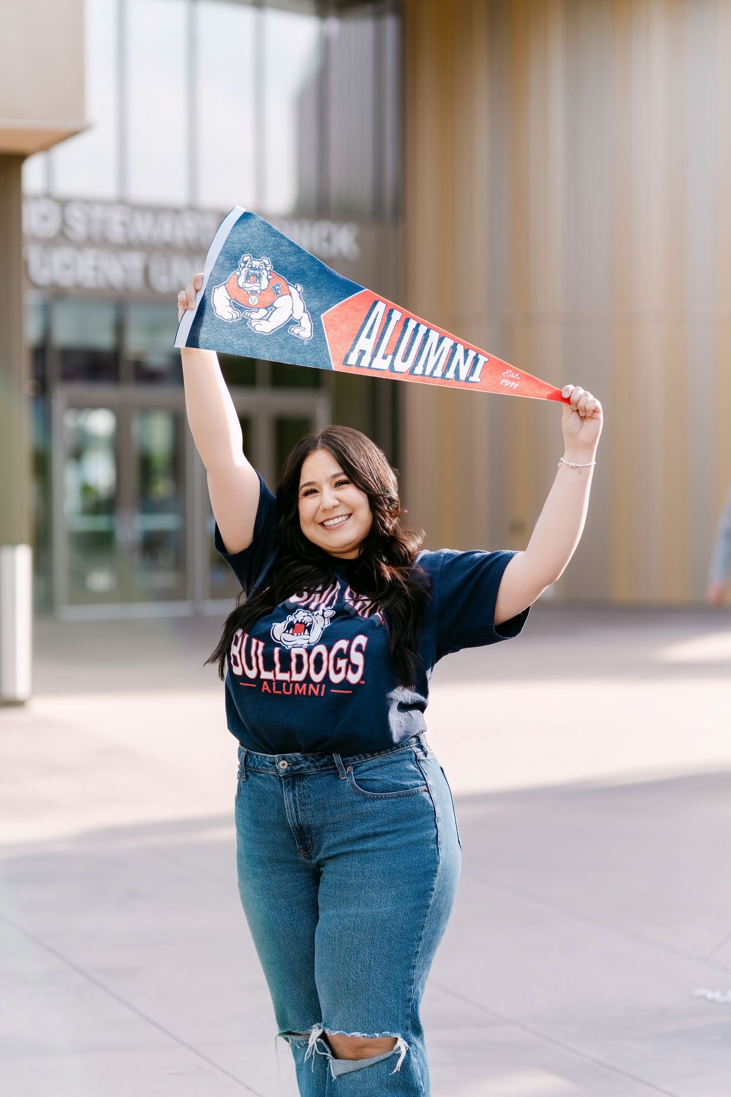 Smiling Fresno State Bulldogs alumni woman holding pennant flag outside university building wearing jeans and Bulldogs shirt.