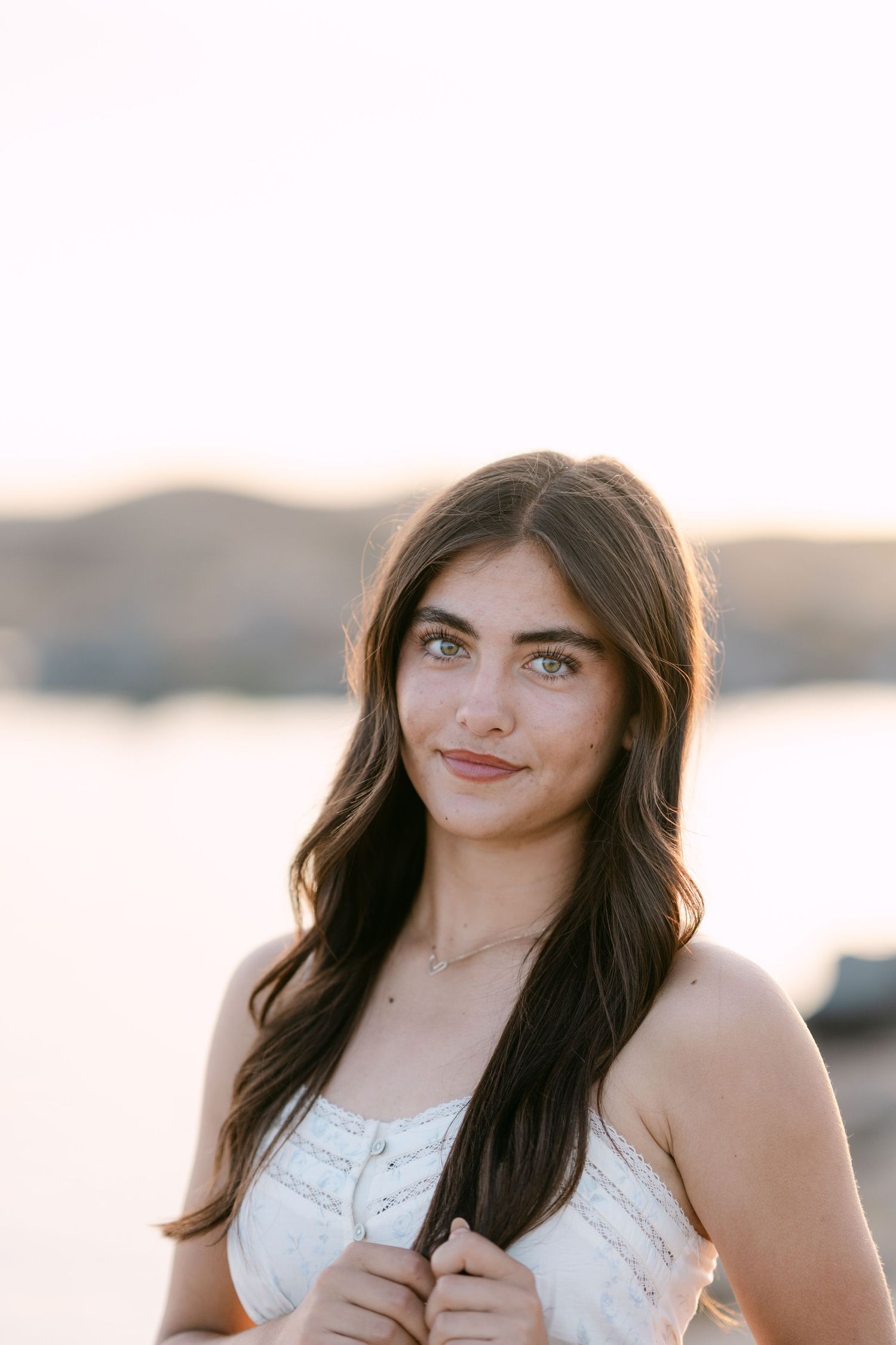Young woman with long dark hair and blue eyes wearing a white top, posing outdoors at golden hour near a lake.