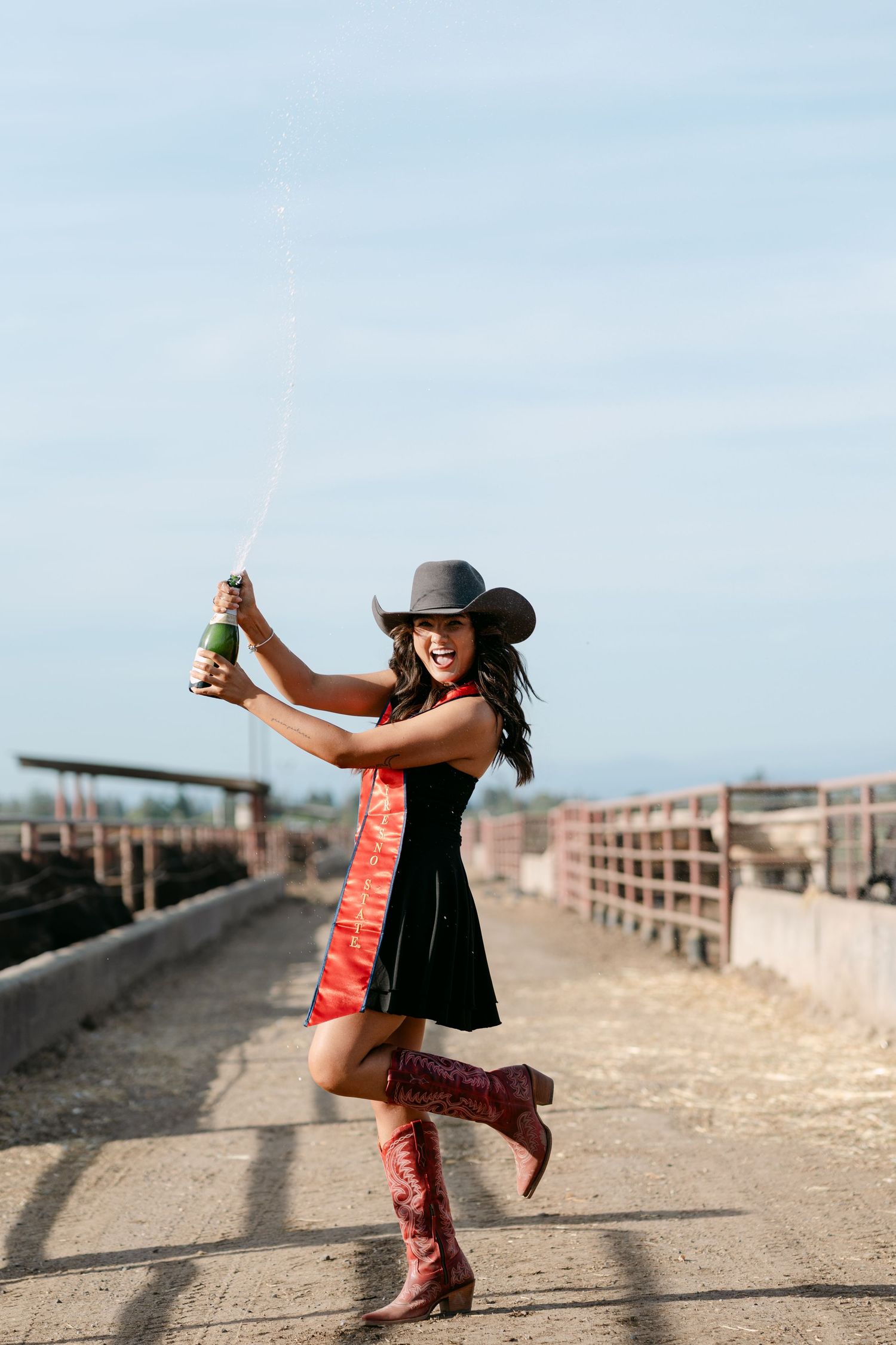 Woman in black dress, cowboy hat, and red boots sabering a champagne bottle outdoors.