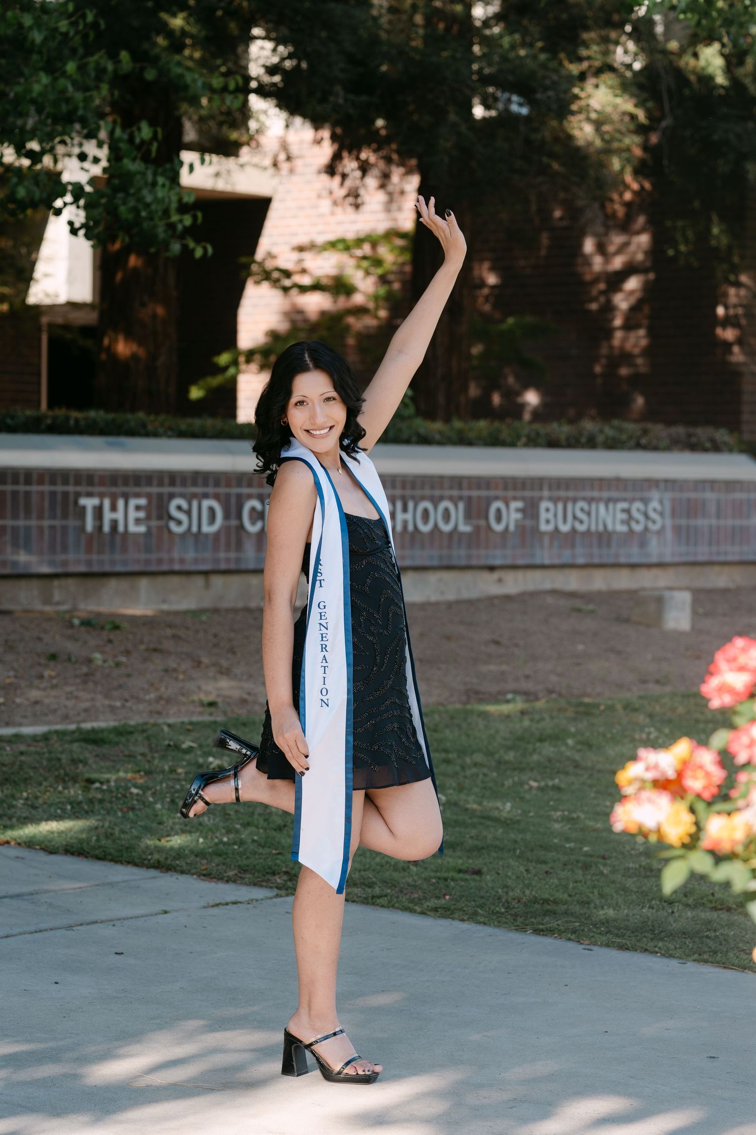 Graduate poses joyfully in front of Sid Craig School of Business wearing a white sash and black dress.
