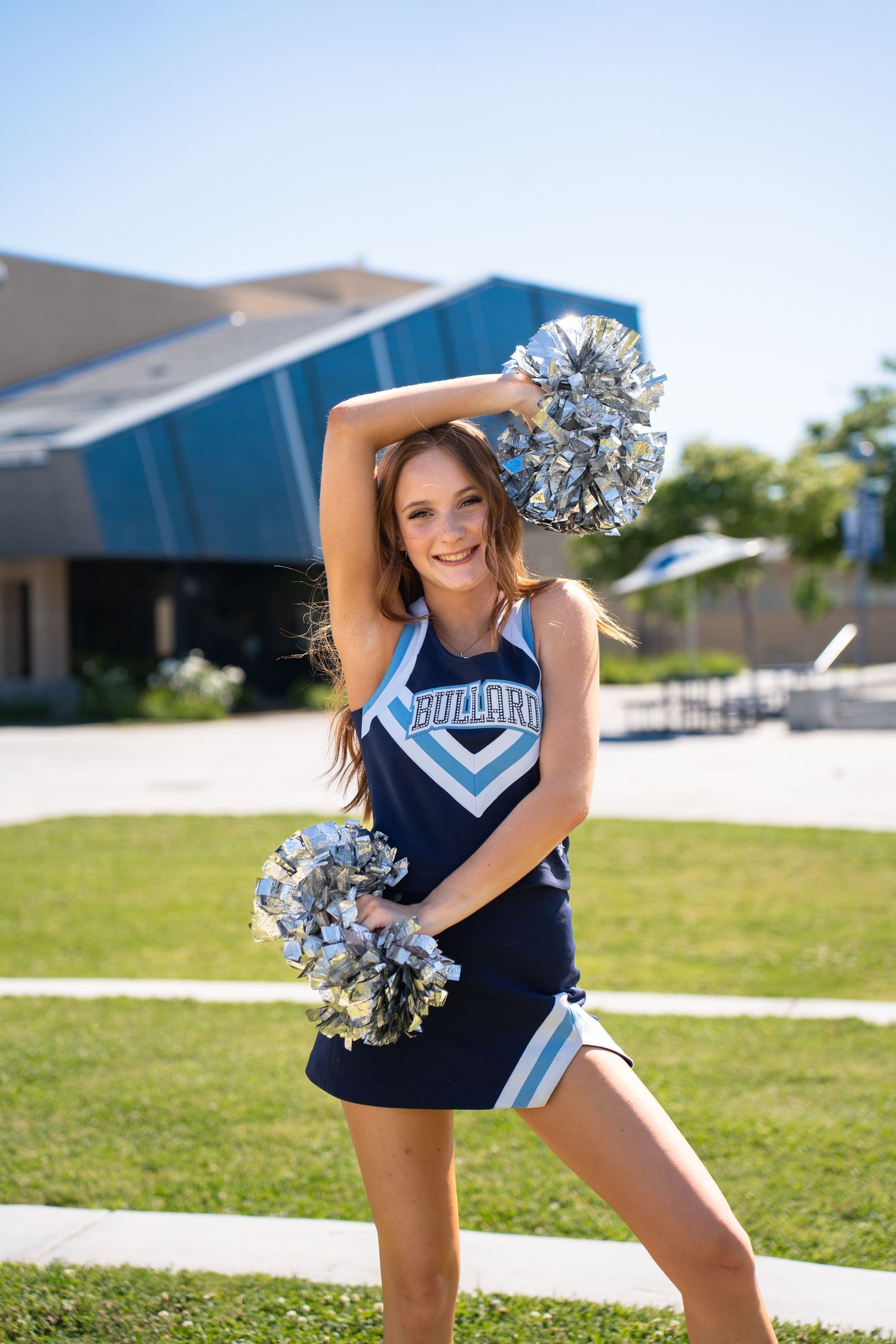 Cheerleader performs dynamic dance moves with silver pom poms on sunny school grounds.