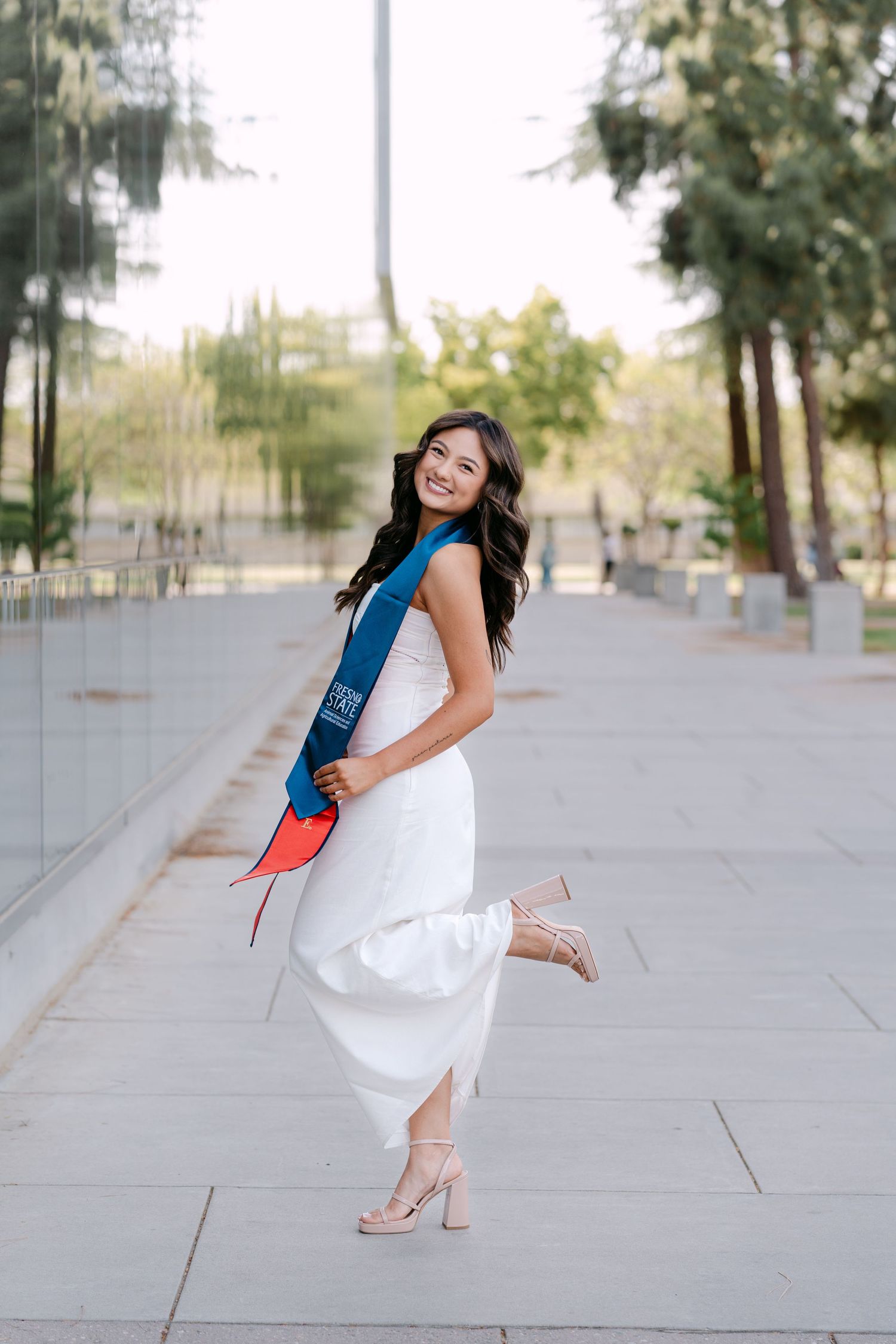 Smiling graduate in white dress and blue sash holds cap while posing outdoors on a tiled walkway.