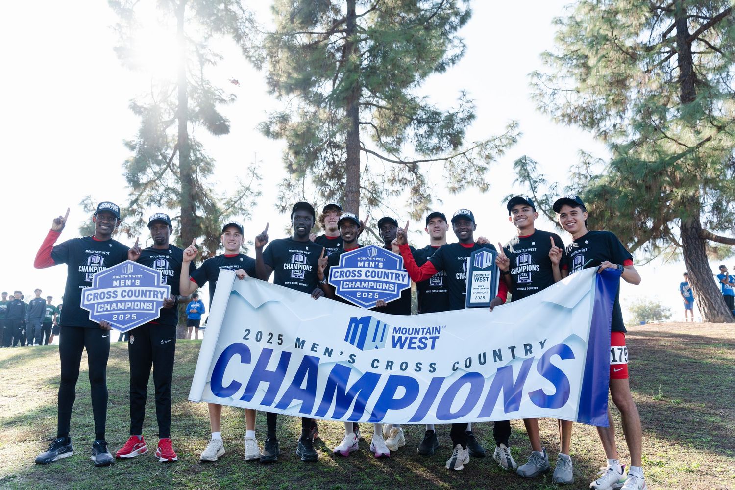 A group of athletes holding a large blue championship banner outdoors on a grassy hillside with pine trees.