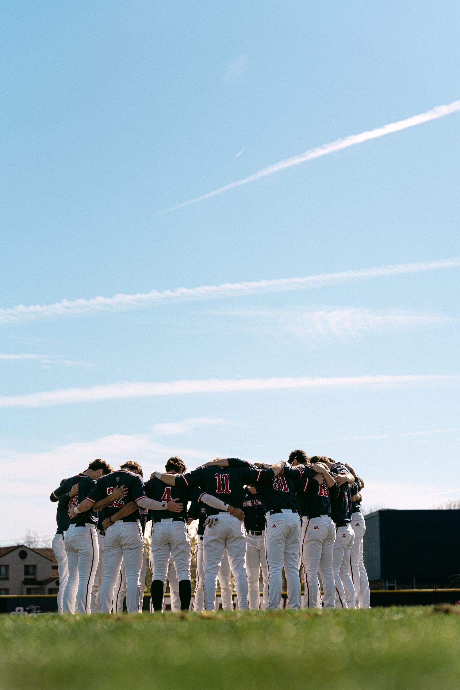 Baseball team huddles together on field against blue sky with contrails visible overhead.