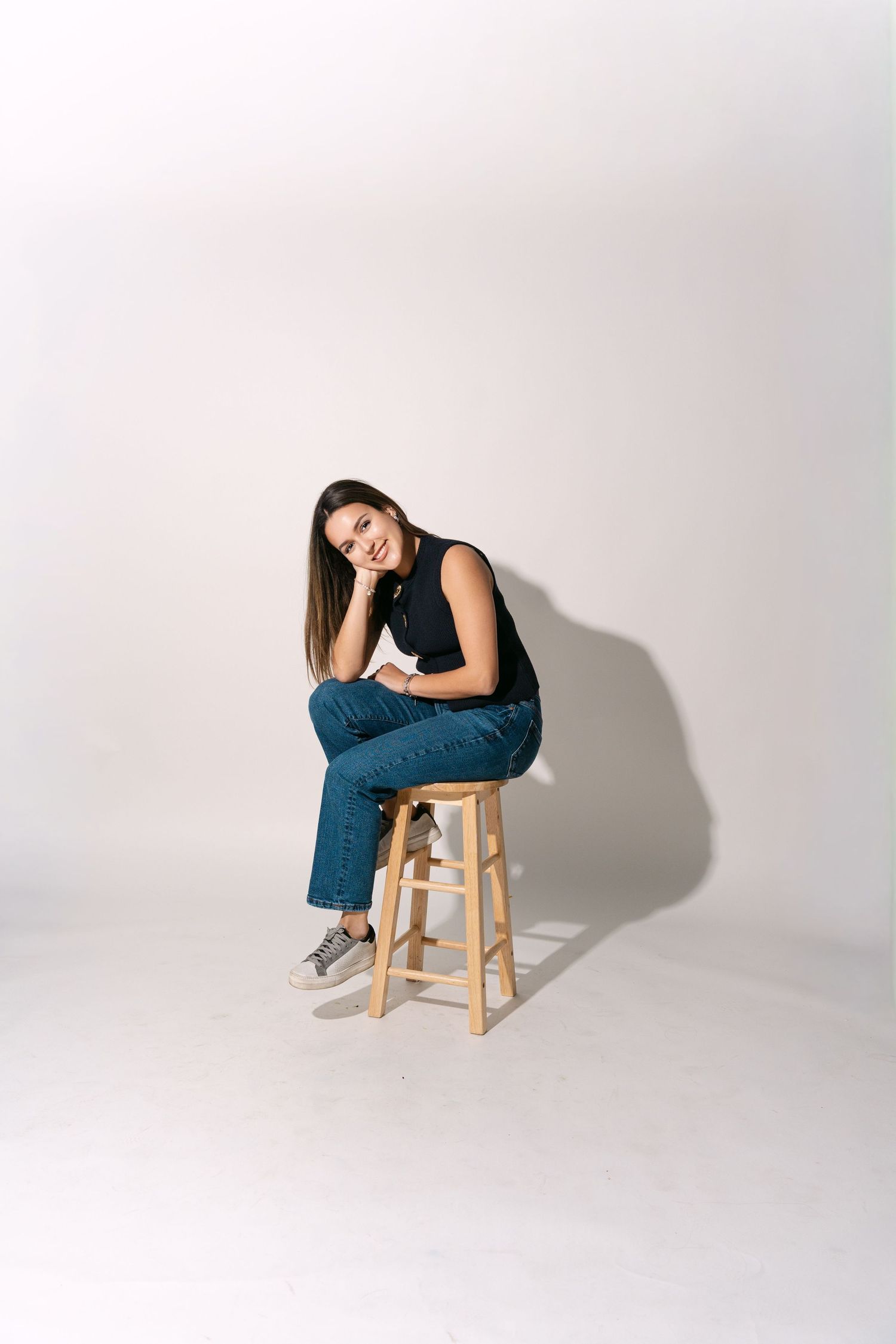 Smiling woman in black top and jeans sitting on a wooden stool against a white background.